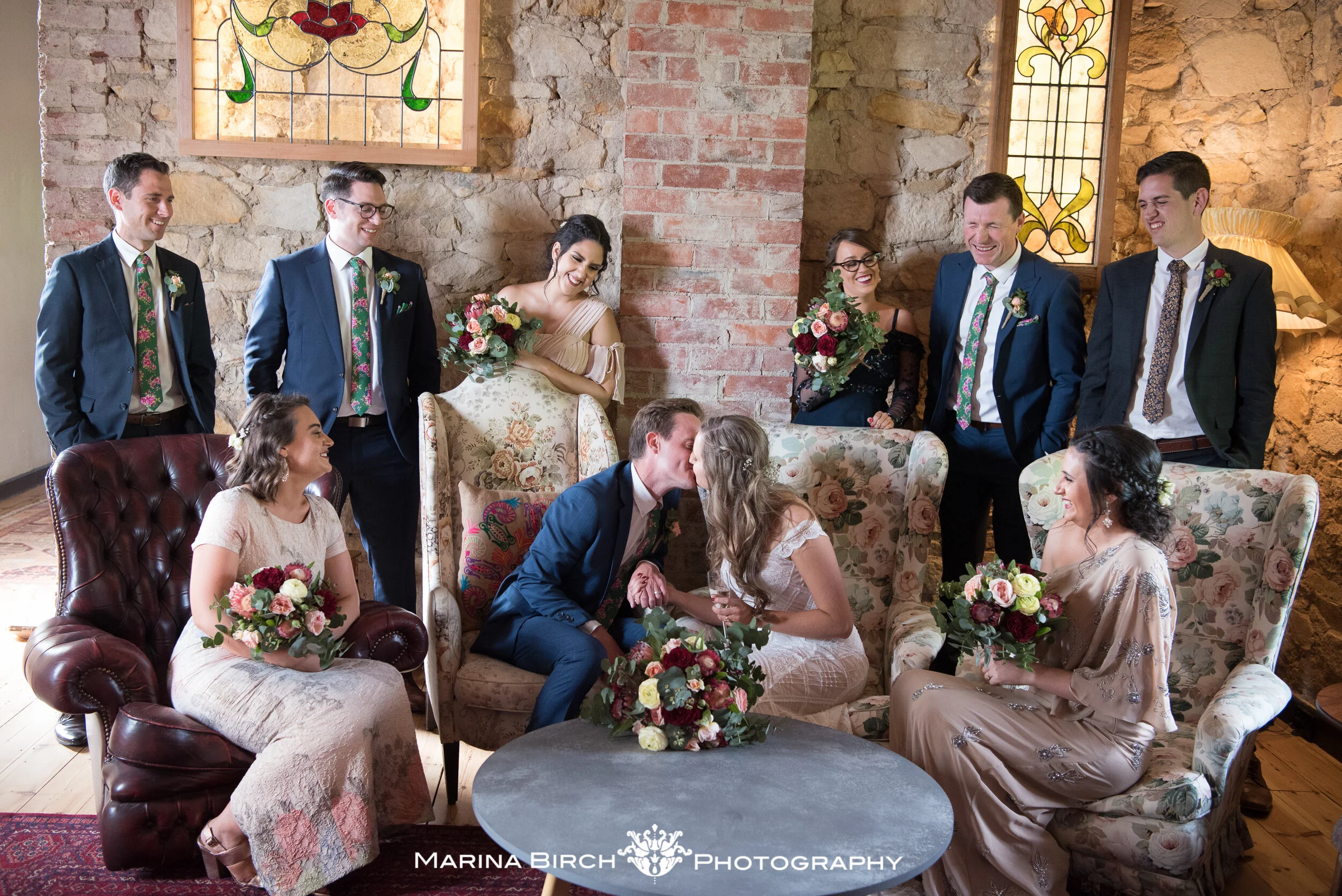 A wedding ceremony with the bride and groom kissing while seated on a floral sofa, surrounded by bridesmaids and groomsmen in a rustic room with stone walls and stained glass windows, holding bouquets of flowers.