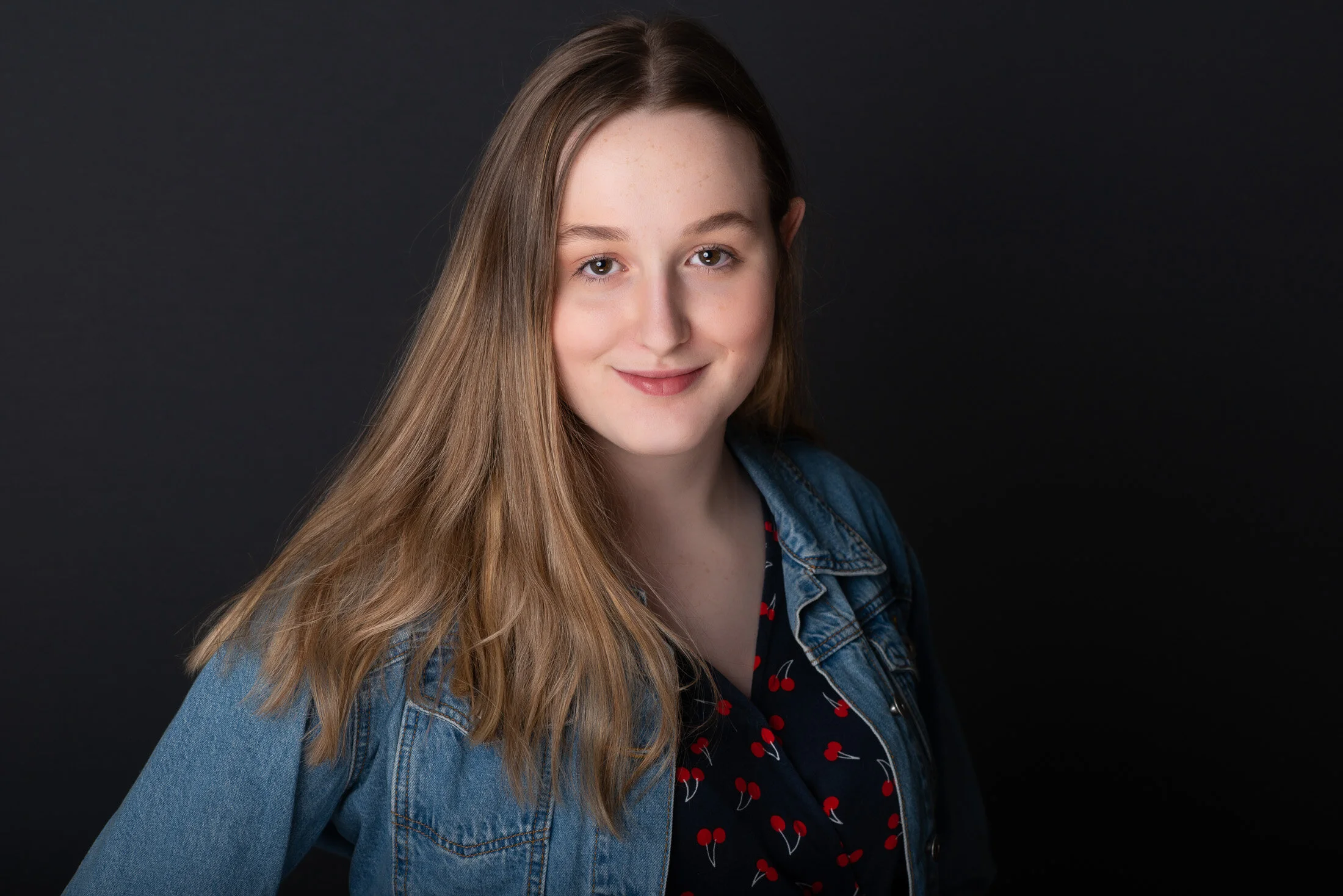 Portrait of a young woman with long light brown hair, wearing a denim jacket and a dark dress with a cherry pattern, against a black background.