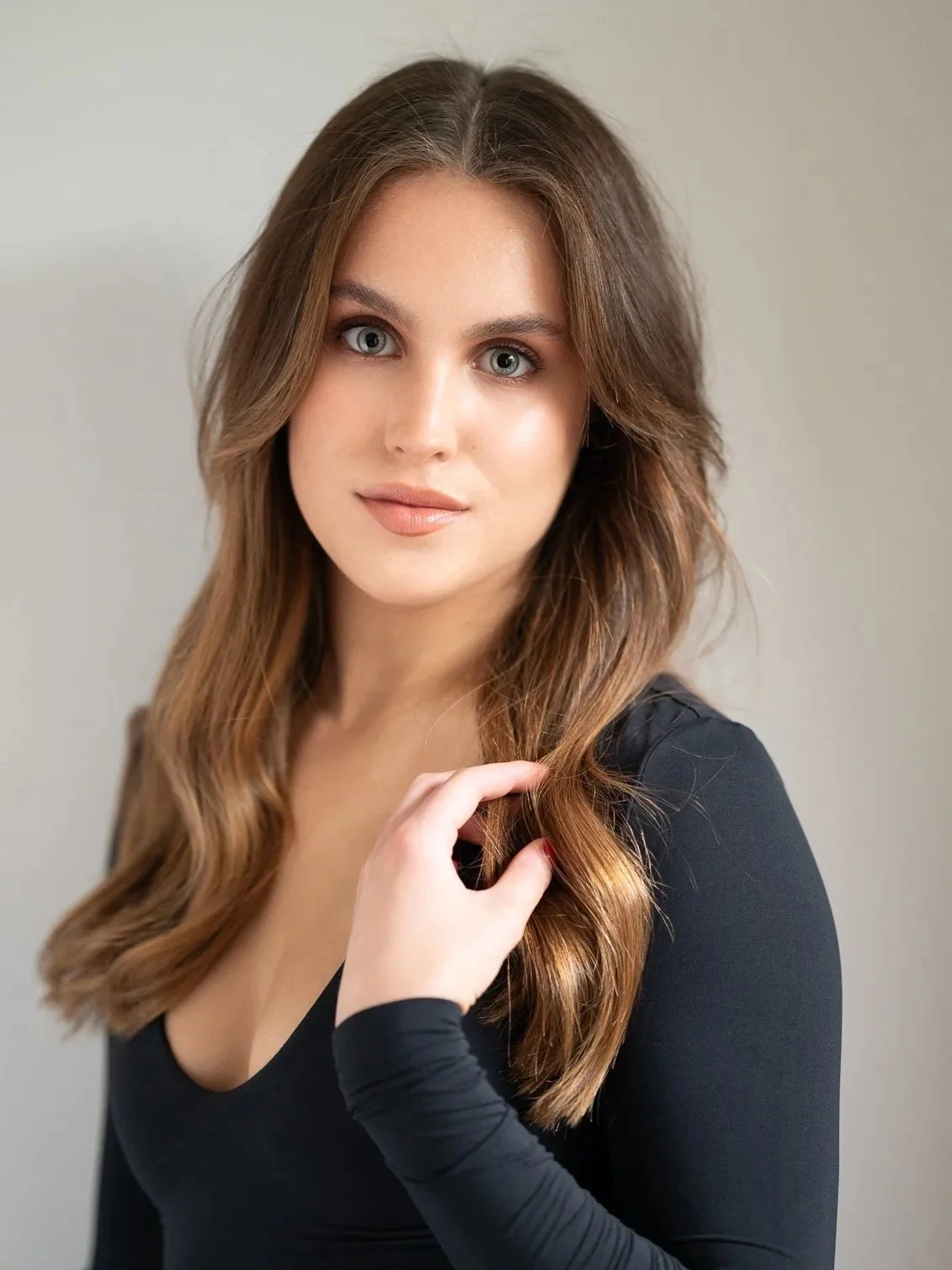 A young woman with long, wavy brown hair, blue eyes, and fair skin, wearing a black top, touching her hair with one hand, looking at the camera with a slight smile.