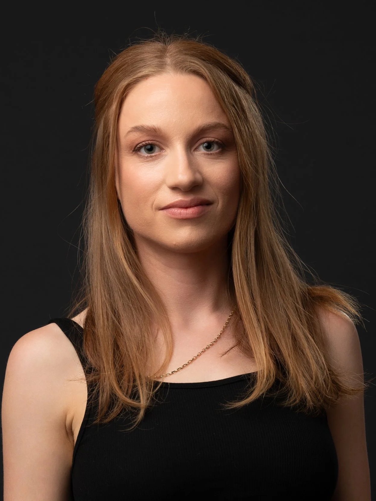 A portrait of a young woman with wavy auburn hair, blue eyes, wearing a black sleeveless top and a gold chain necklace, against a black background.