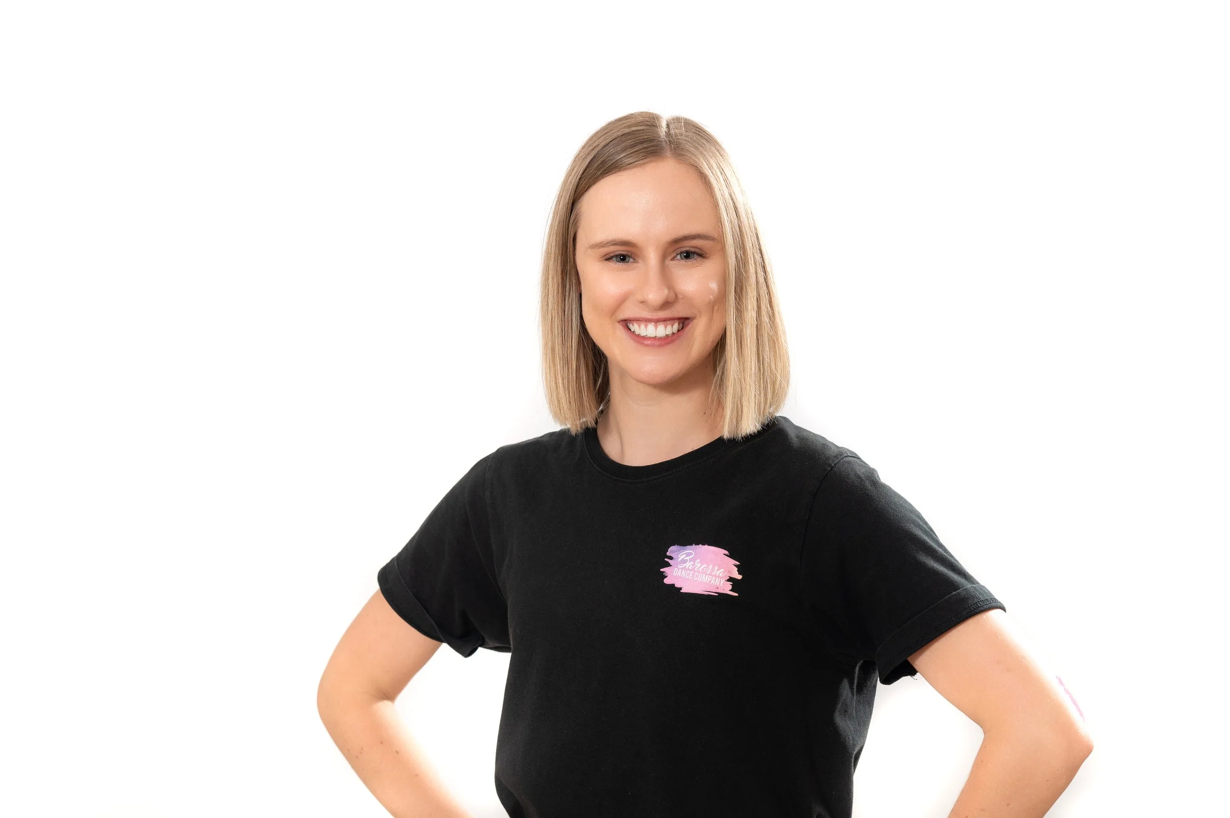 A young woman with shoulder-length blonde hair smiling and posing with her hands on her hips, wearing a black t-shirt with a pink and purple graphic logo on the chest against a plain white background.Adelaide dance school photography
Photoday