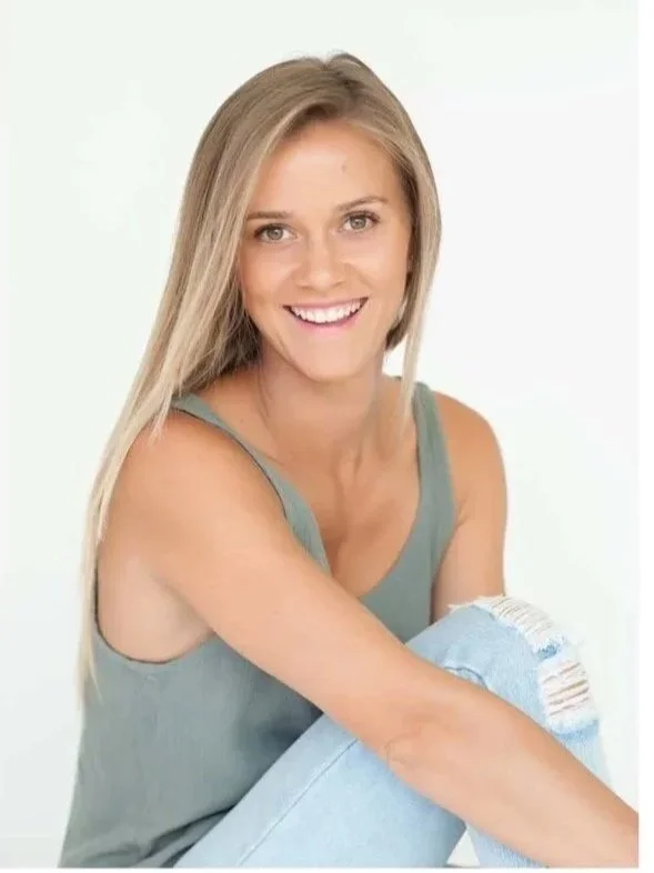 A woman with long blonde hair smiling, wearing a sleeveless gray top and ripped jeans, sitting against a plain white background.