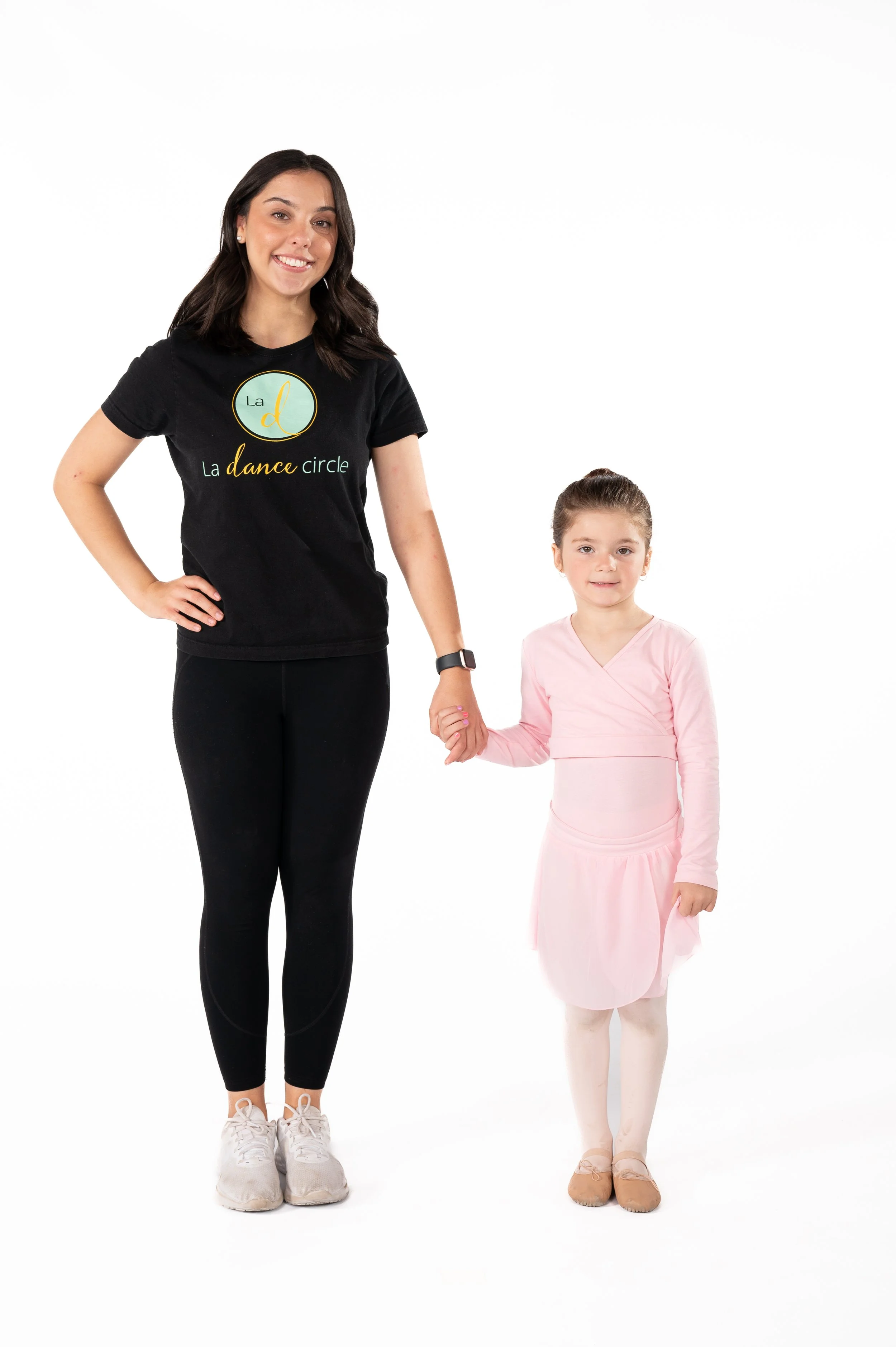 A young adult woman in a black t-shirt and black leggings standing next to a young girl in a pink ballet outfit, holding hands. The woman is smiling and the girl has a neutral expression.