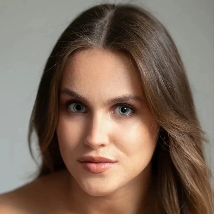 Close-up image of a woman with light skin, brown hair, and blue eyes, looking directly at the camera.