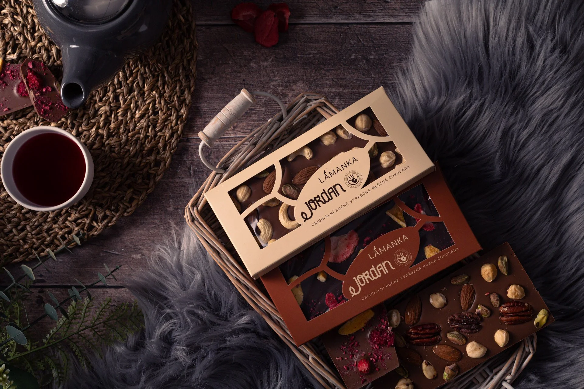 Assorted chocolate bars with nuts and dried fruit in a wicker basket next to a cup of red wine and a teapot on a dark wooden surface with a gray furry blanket and some greenery.  Photo by lucie lizlerova