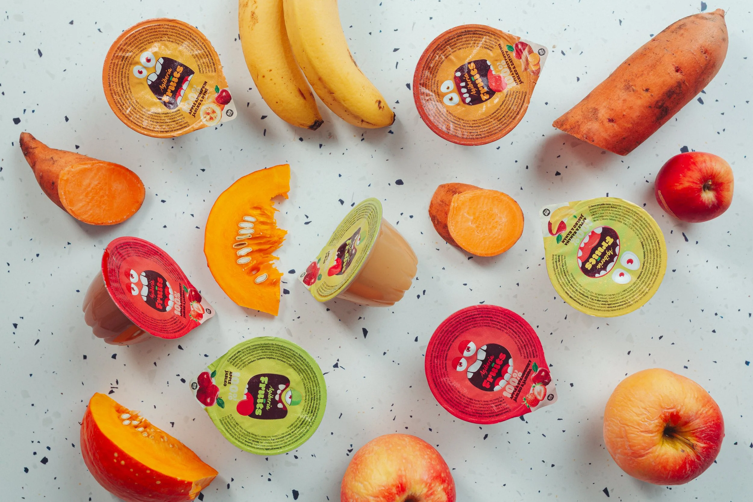 Assorted fruit-flavored jelly cups, whole bananas, sweet potatoes, apples, sliced pumpkin, and peeled sweet potato on a speckled white countertop.  Photo by lucie lizlerova