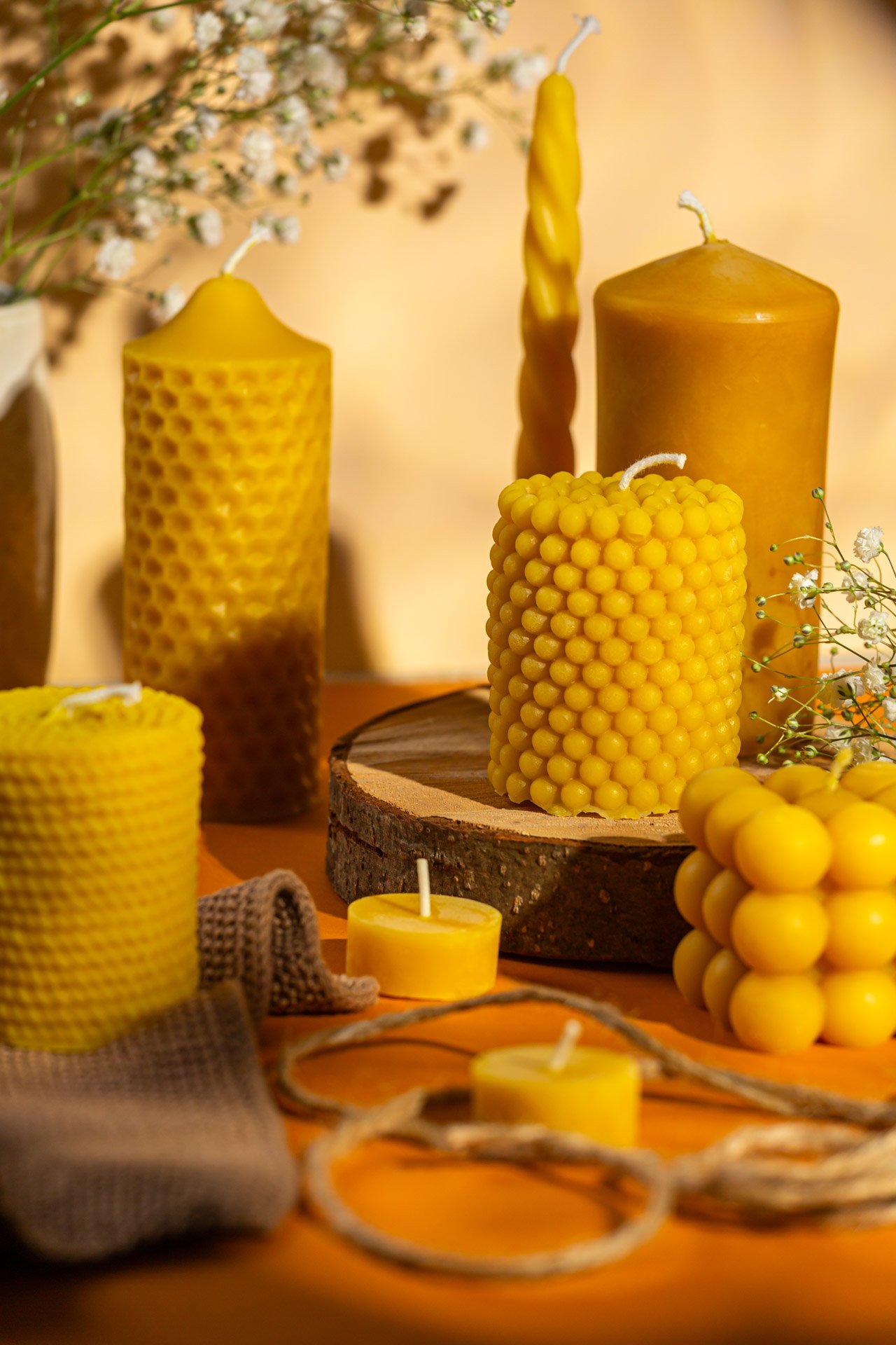 Yellow and brown candles of various shapes and sizes arranged on a wooden surface with some decorative dried flowers and twisted cords.  Photo by lucie lizlerova