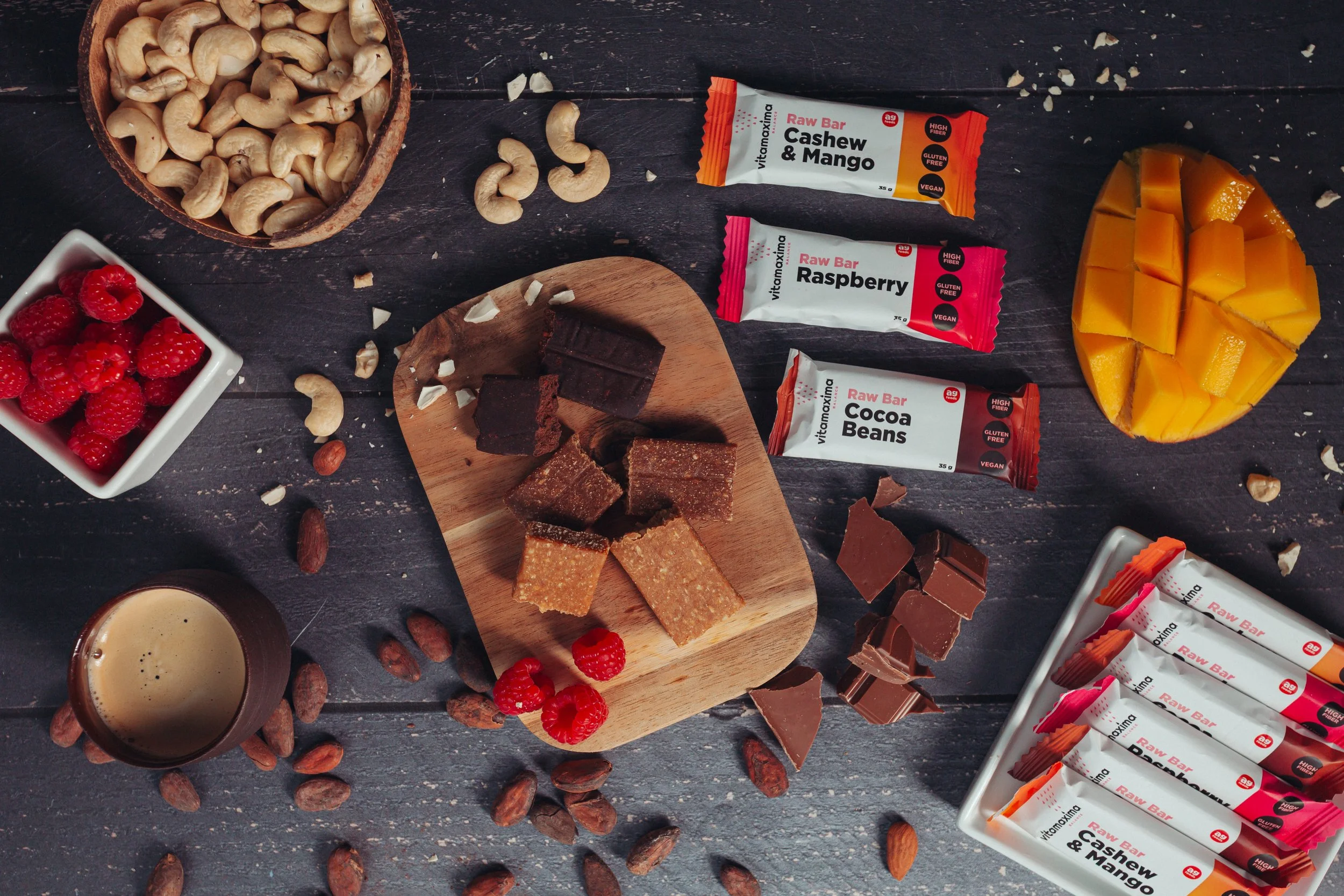 A flat lay of various snacks and ingredients on a dark wooden surface, including bowls of cashews and raspberries, a chocolate and fruit smoothie, honeycomb, chocolate bars, raw energy bars labeled cashew & mango, raspberry.  Photo by lucie lizlerova