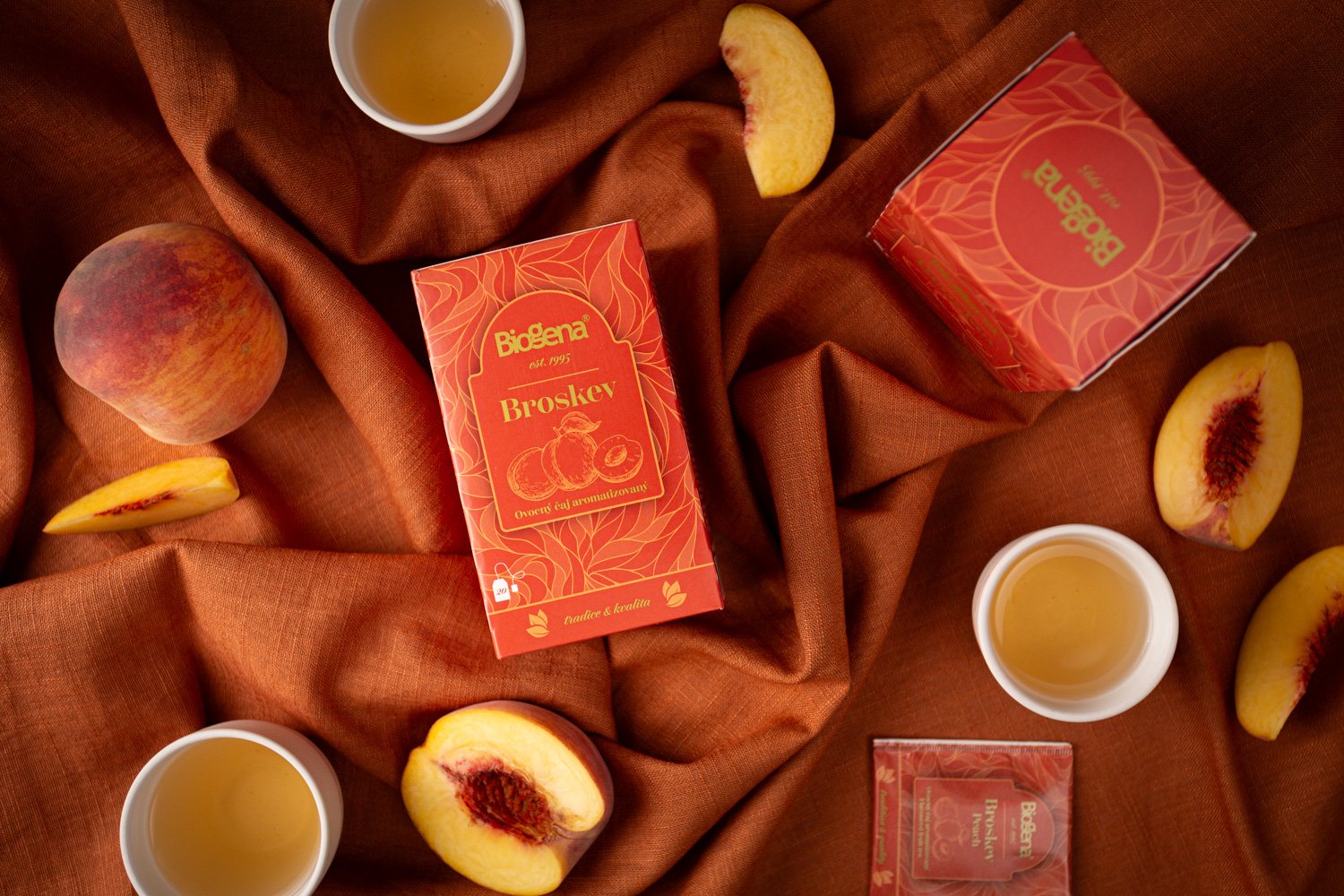 Box of Biogena Broskev peach flavored fruit tea, sliced peaches, peach tea in cups, and peach slices on a brown fabric background.  Photo by lucie lizlerova