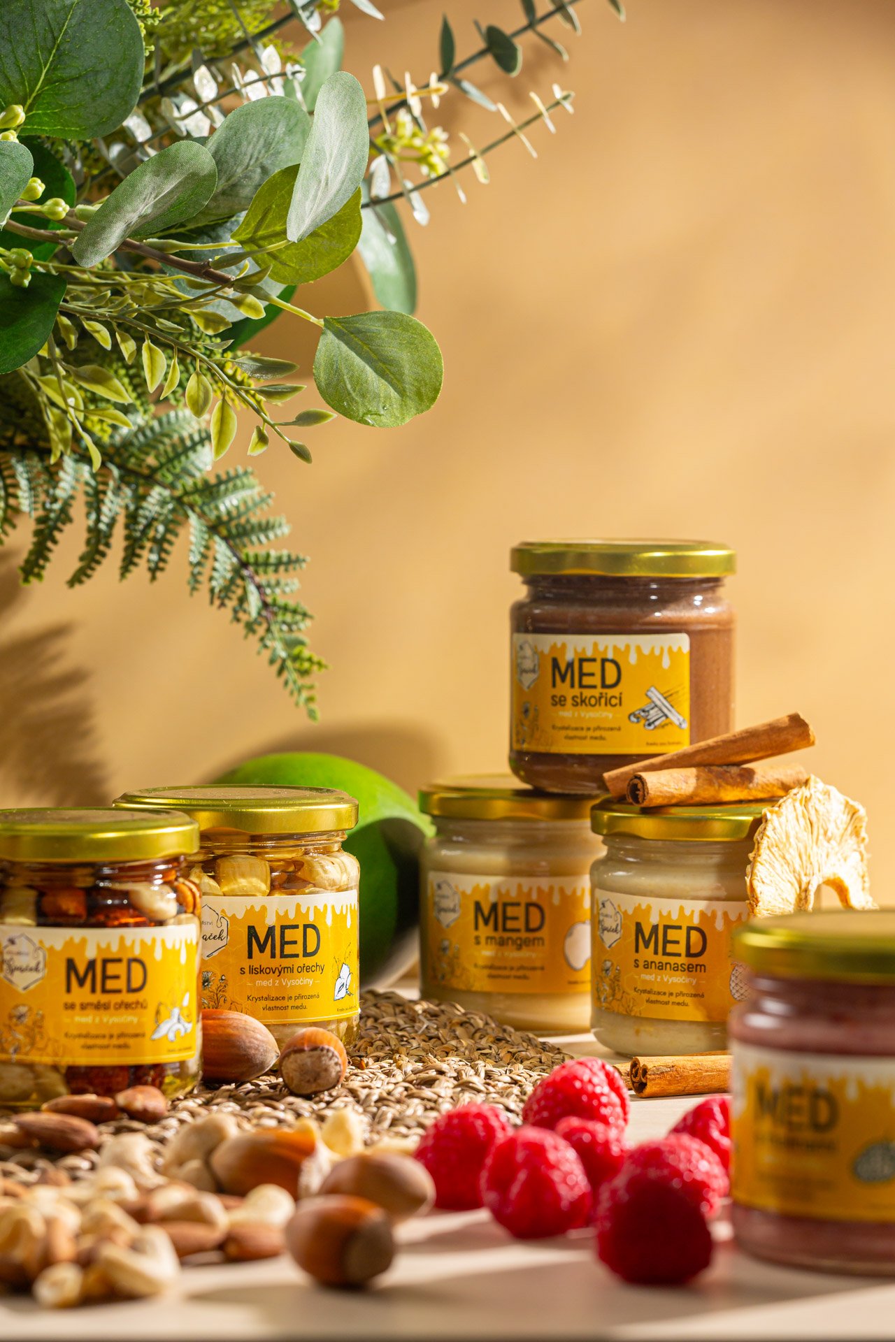 Assorted jars of honey with honeycombs and dried fruit, surrounded by nuts, raspberries, apple, cinnamon sticks, and green foliage on a light background.  Photo by lucie lizlerova