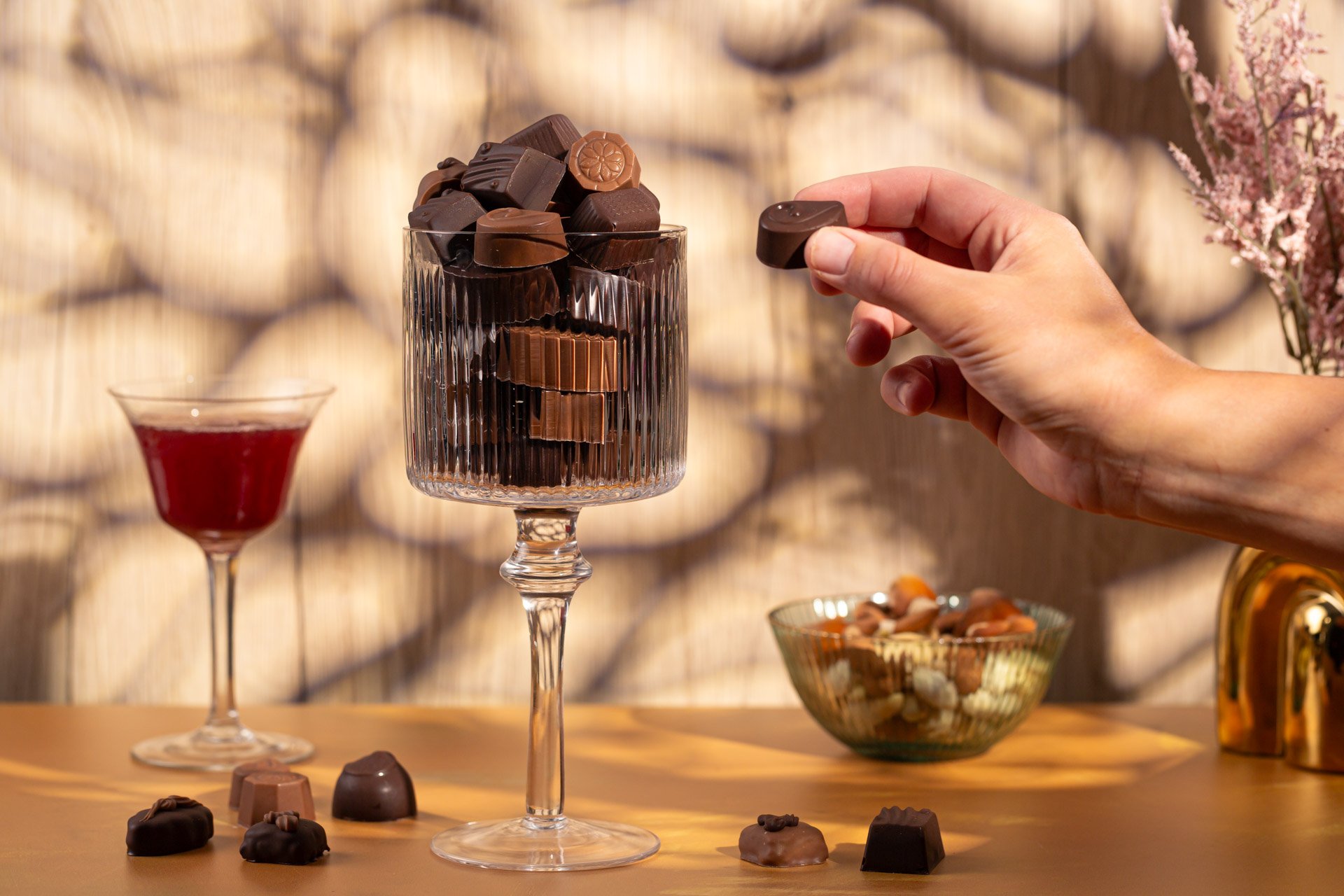 A hand placing a piece of chocolate into a glass filled with assorted chocolates and chocolates inside a glass container. A bowl of mixed nuts and a glass of red wine are on the table in the background.  Photo by lucie lizlerova