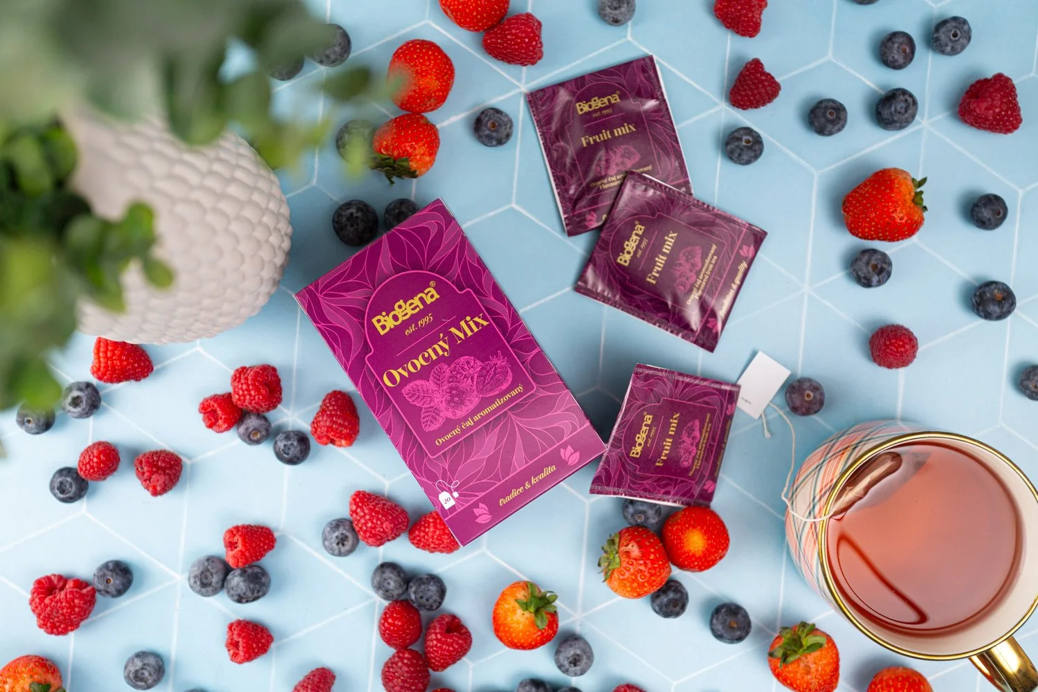 A box and three sachets of Biogena fruit mix tea, scattered strawberries and blueberries on a light blue tiled surface, with a white textured plant pot on the left and a cup of brewed tea or coffee on the right.  Photo by lucie lizlerova