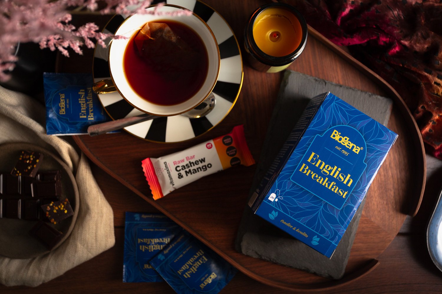 A breakfast setup on a wooden table includes a blue box of Brogena English Breakfast tea, a cup of dark tea on a black-and-white striped saucer, a yellow candle in a jar, a blue cashew and mango snack bar, a small bowl of chocolates with colorful spr