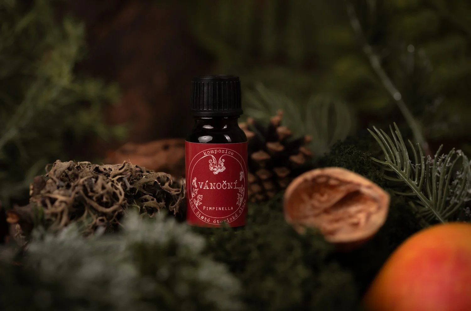 A small dark bottle with a red label surrounded by pine cones, dried orange slices, and evergreen branches.  Photo by lucie lizlerova