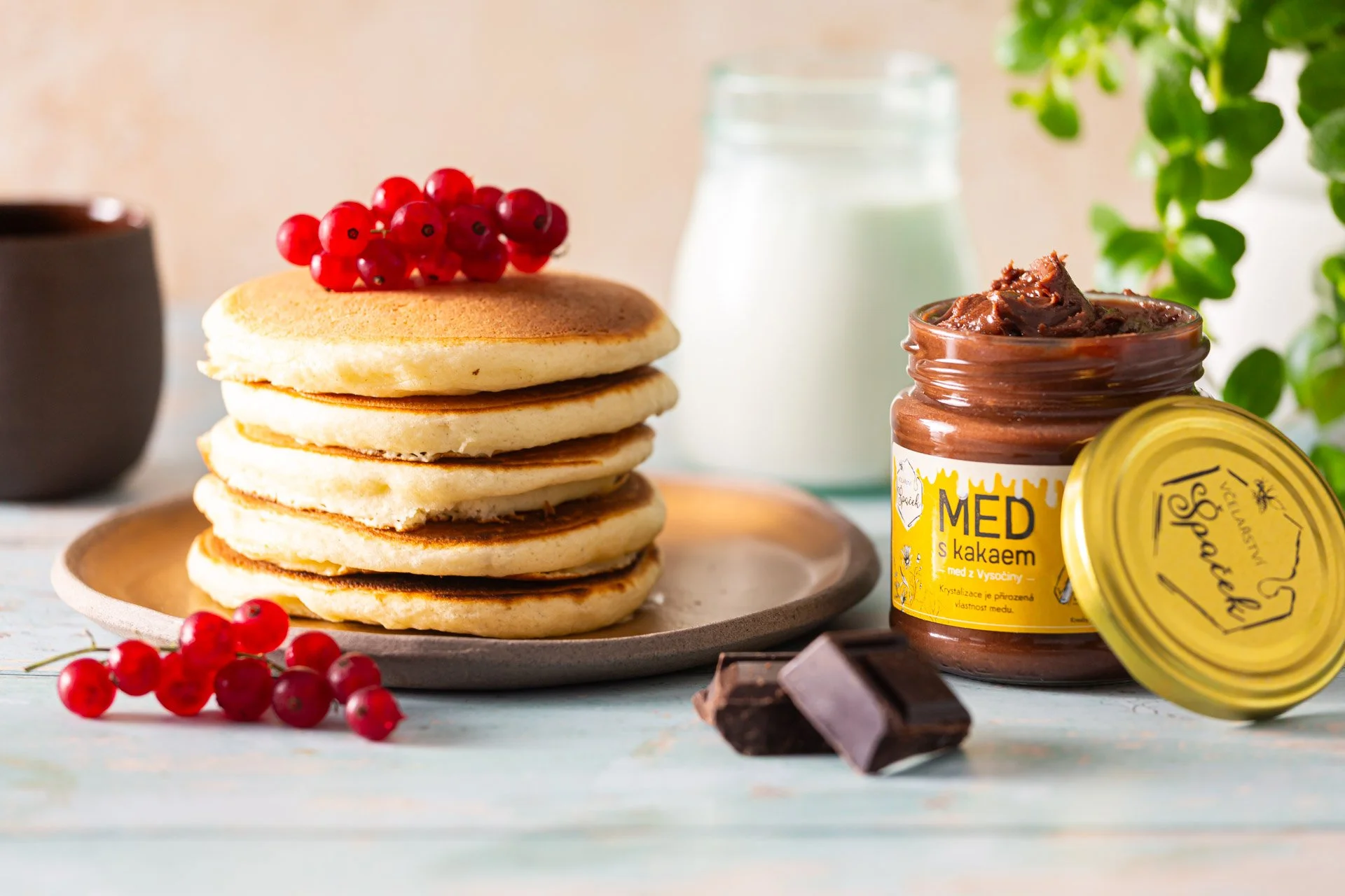 Stack of five pancakes topped with red currants, with a jar of honey with cocoa, a chocolate piece, a glass bottle of milk, a black coffee mug, and green plants in the background. Photo by lucie lizlerova
