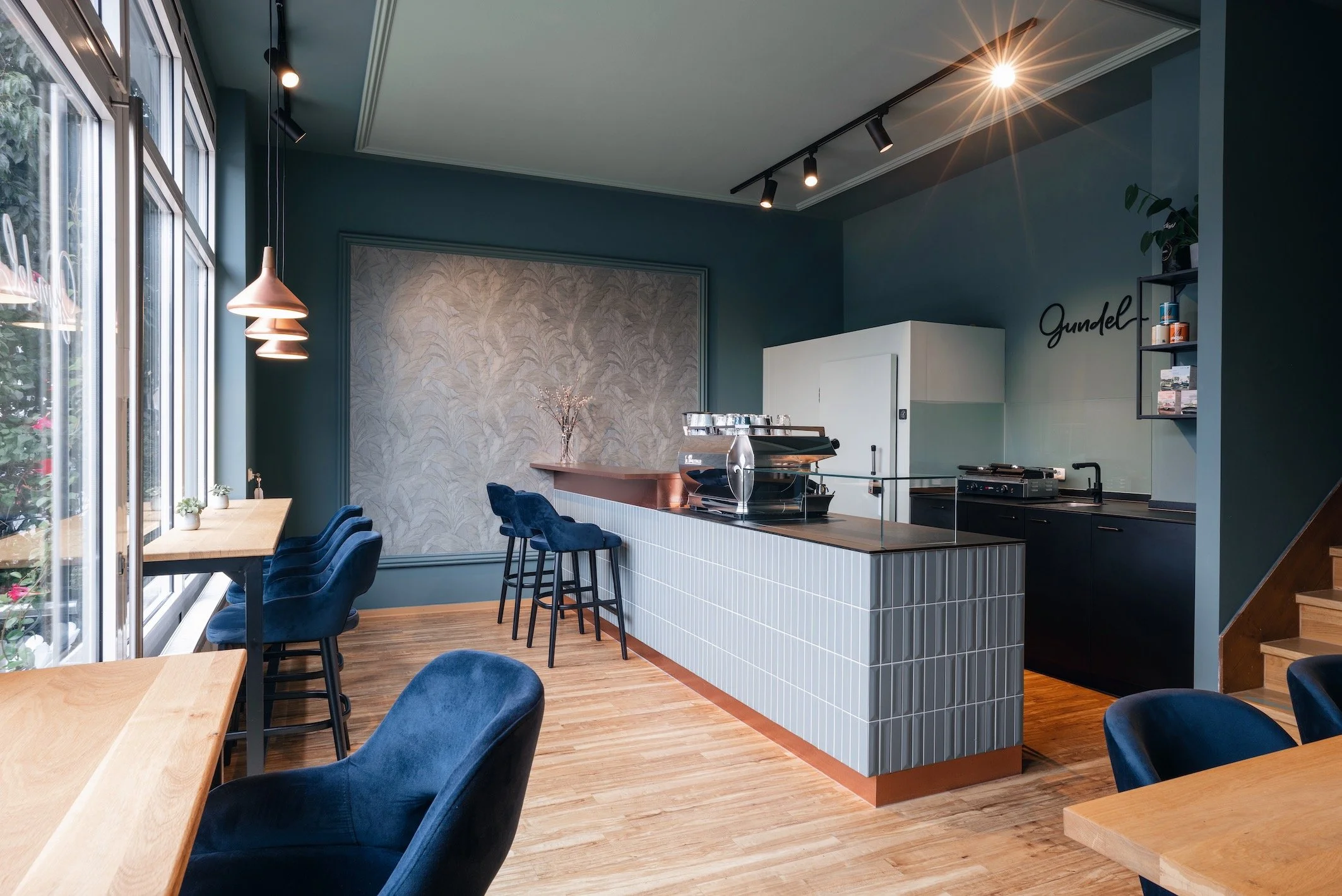 Gemütliches Café mit blauen Stühlen, Holztischen, einer Theke mit grauen Fliesen, einem Kaffeemaschine und gutem natürlichen Licht durch große Fenster.