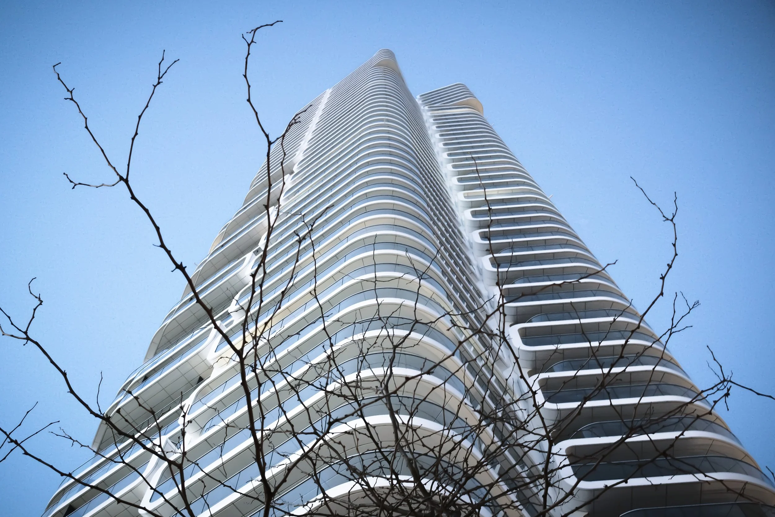 Hoher moderner Wolkenkratzer mit geschwungenen Balkonen, Blick nach oben, im Vordergrund kahle Baumzweige, blauer Himmel