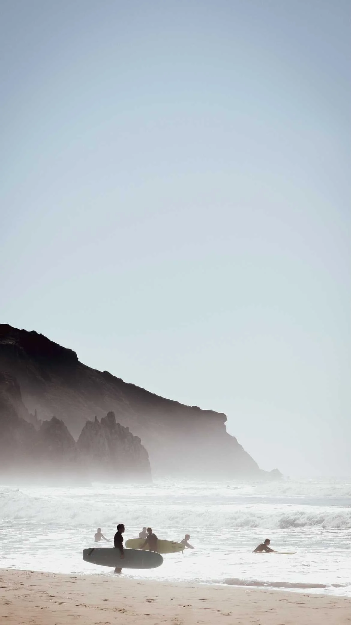 Menschen am Strand mit Surfbrettern, große Klippen im Hintergrund, leicht nebliges Wetter