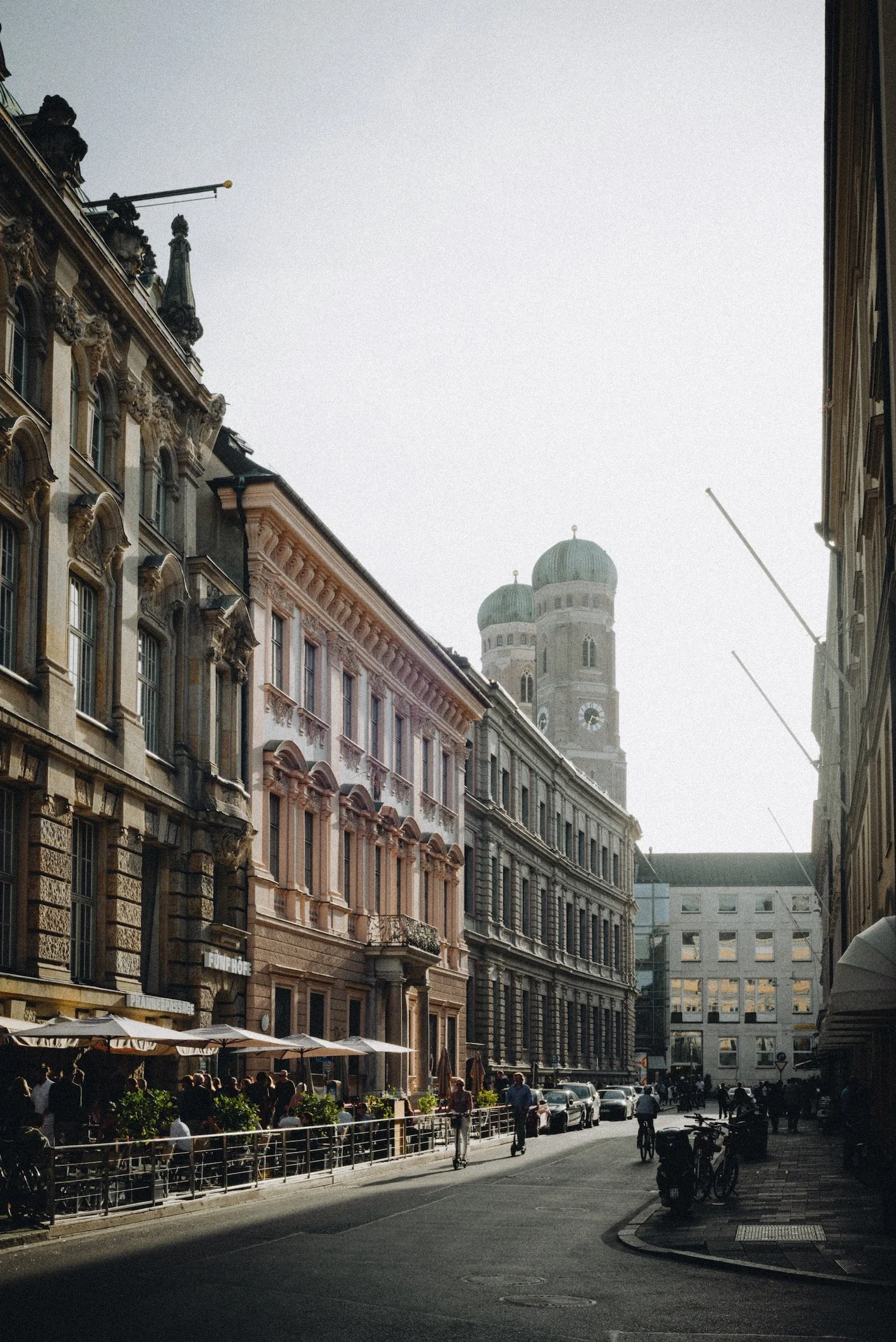 Straße in einer europäischen Stadt mit historischen Gebäuden, Café-Terrassen und Kirchturm im Hintergrund