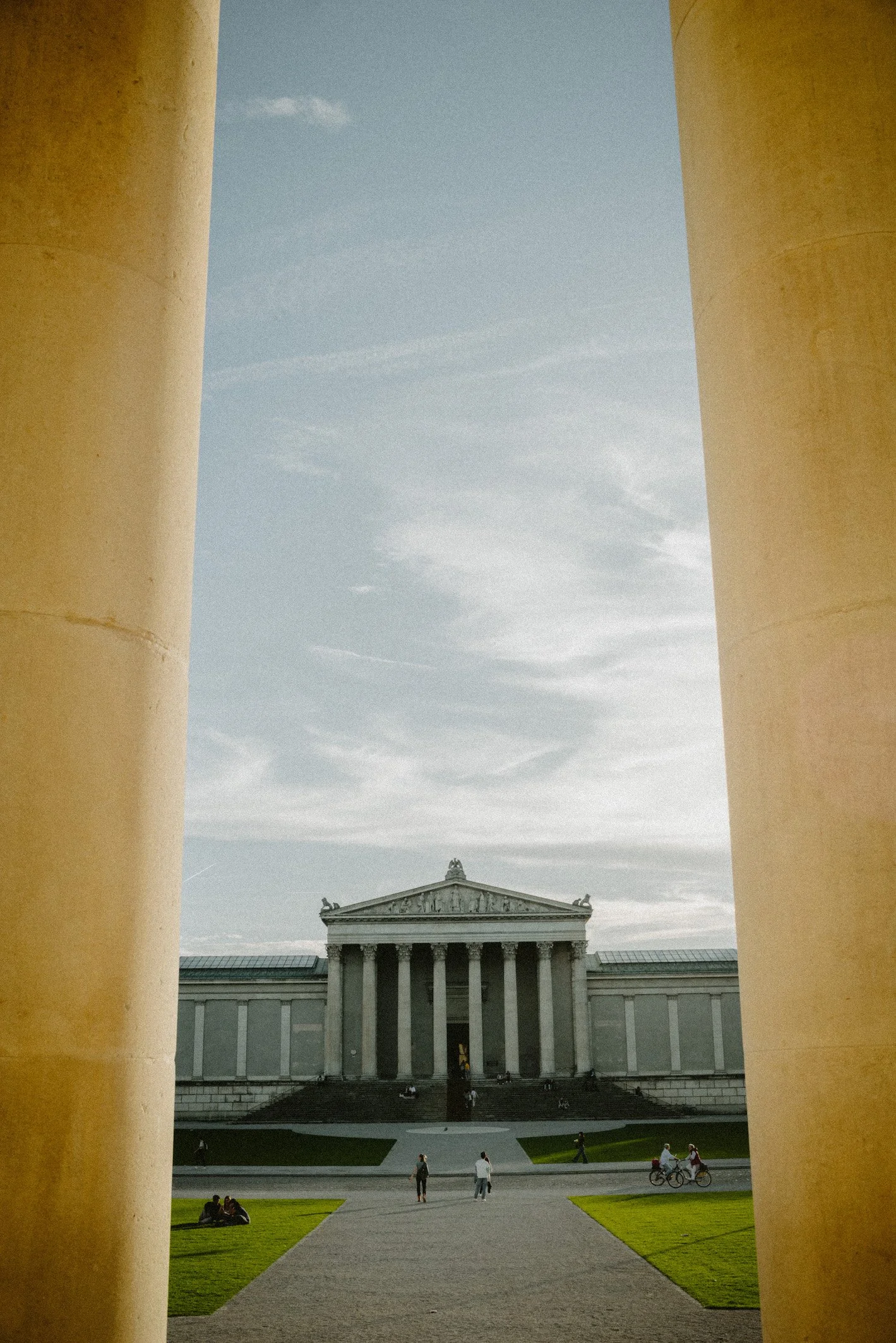 Blick auf ein neoklassizistisches Gebäude mit Säulen, gesehen durch eine Öffnung zwischen zwei großen Säulen, mit Menschen im Vordergrund und einem Himmel mit Wolken.