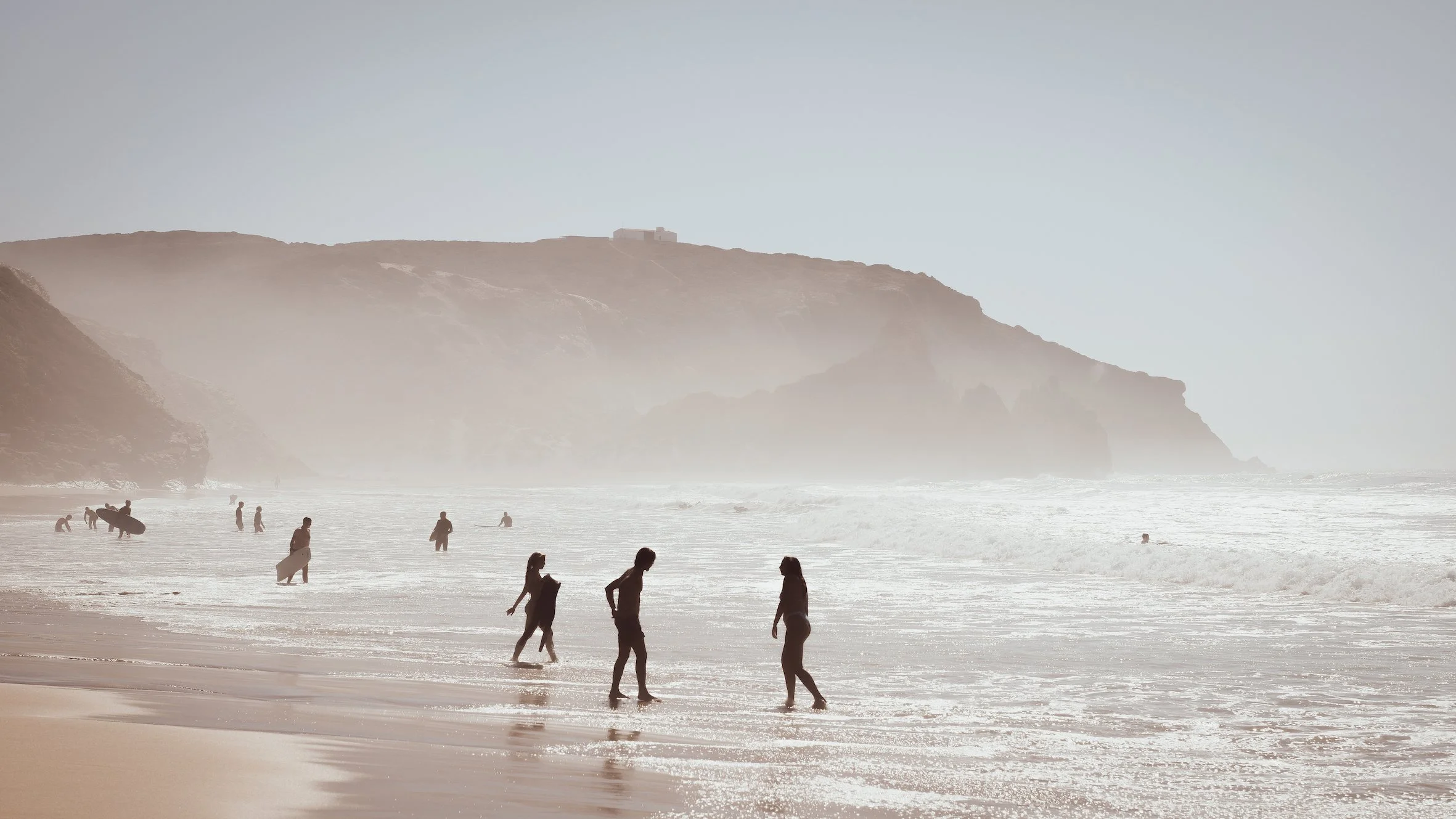 Menschen am Strand beim Spazieren und Surfen, mit Klippen im Hintergrund und atmosphärischem Himmel