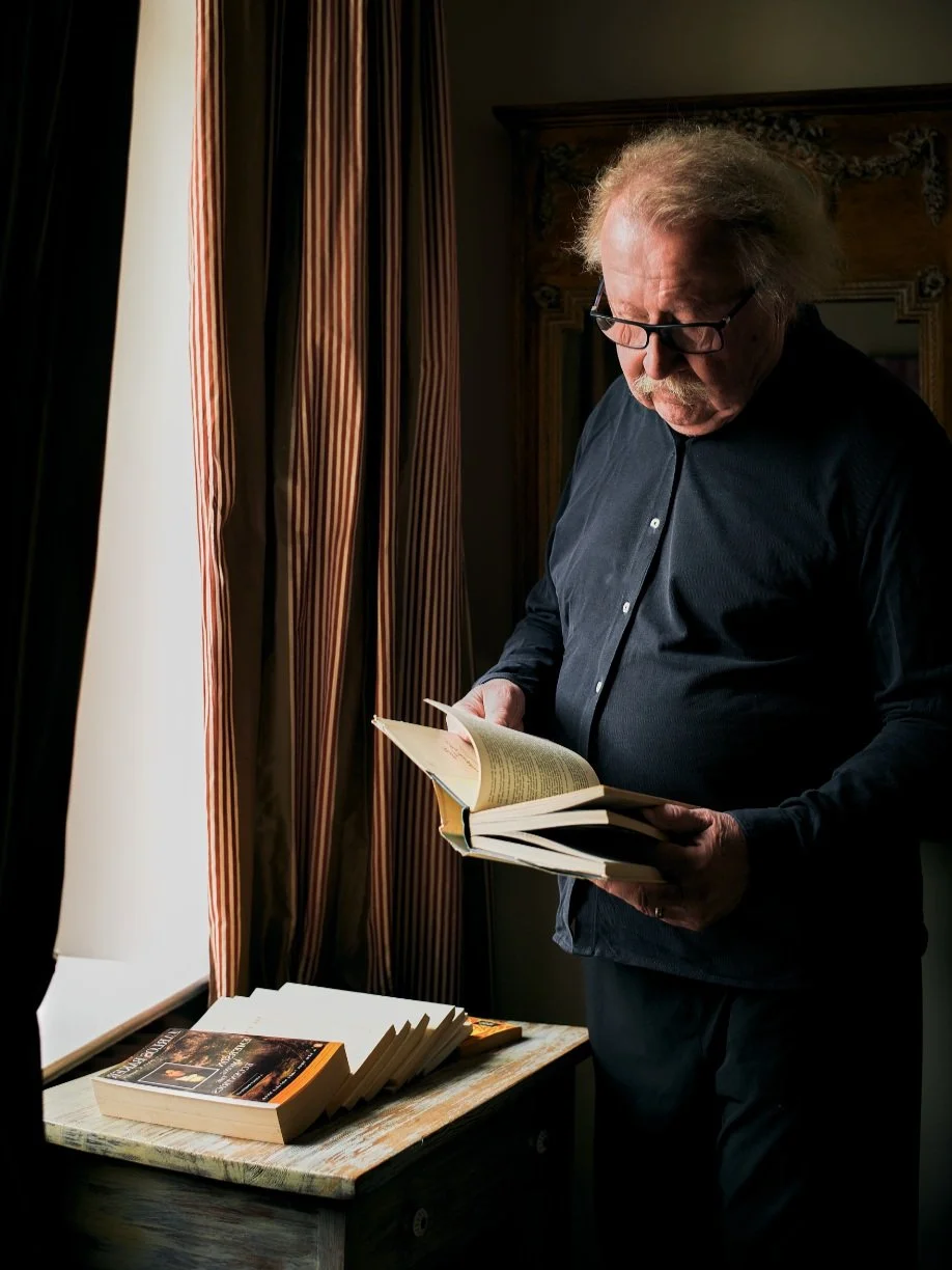 Philiosoph Peter Sloterdijk mit Brille, schwarzem Hemd und grauem, zerzaustem Haar liest ein Buch in einem dunklen Raum. Neben ihm auf einem Tisch liegen weitere Bücher. Im Hintergrund sind lange, gestreifte Vorhänge.