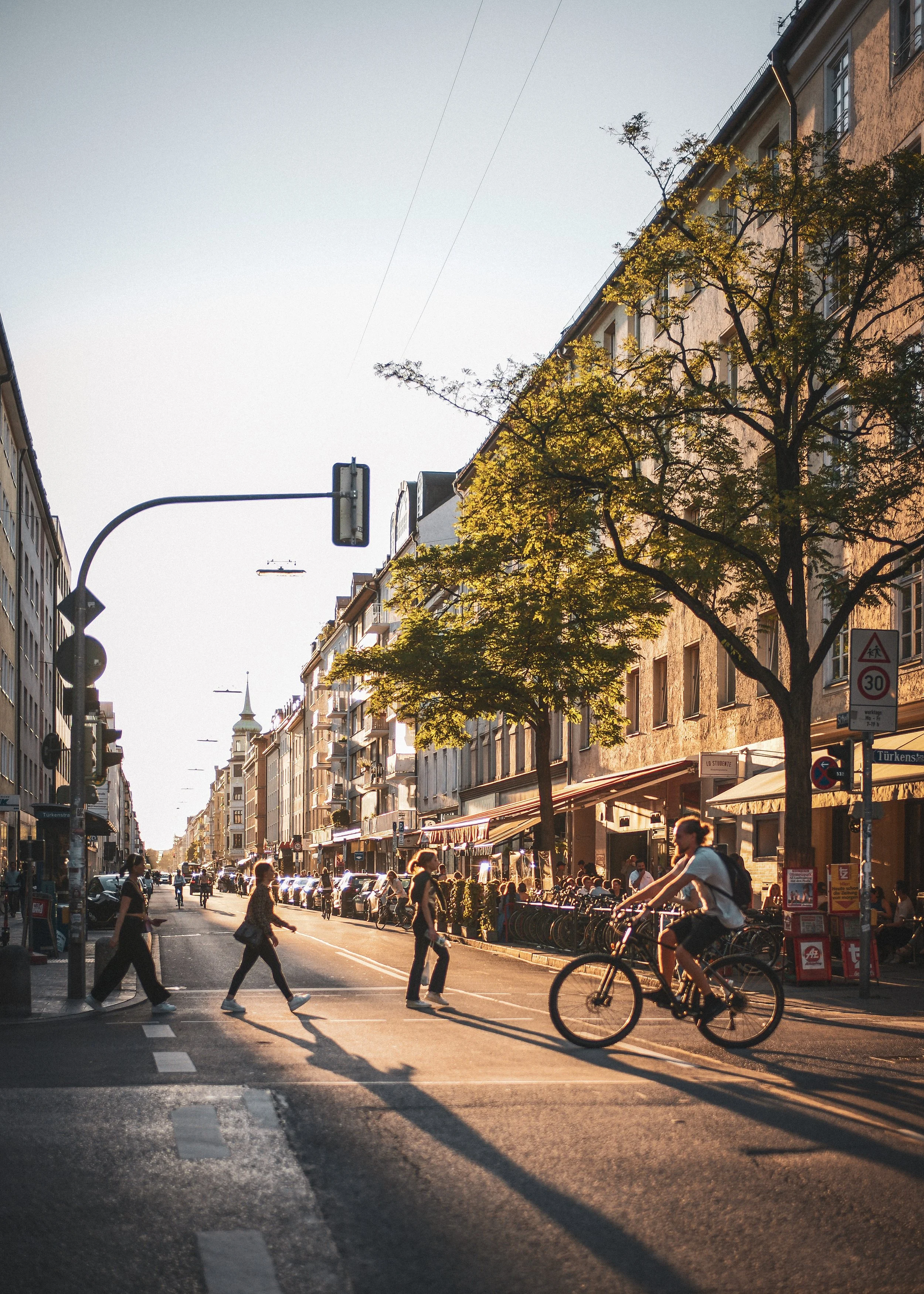 Straße in einer Stadt bei Sonnenuntergang, Menschen getrennt nach Fußgängern und Fahrradfahrern, bunte Gebäude, Bäume, Cafés und Geschäfte an der Seite.