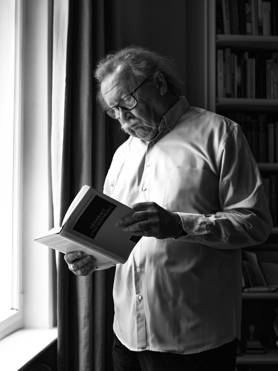 Philiosoph Peter Sloterdijk mit Brille, der ein Buch liest, während er am Fenster steht. Im Hintergrund Bücherregal.