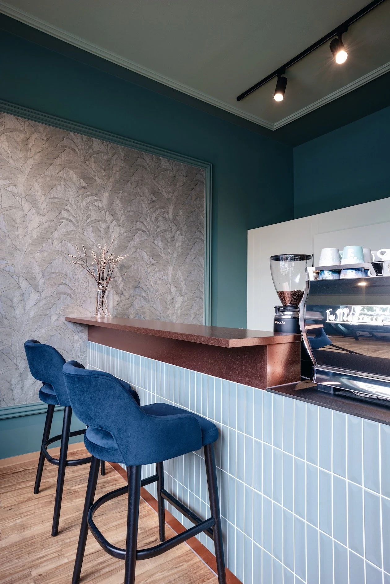 Blick auf eine moderne Café-Theke mit blauen Barhockern, braunem Thekenplatte, weiß geteiltem Fliesenspiegel, eine Kaffee- und Espressomaschine sowie eine Vase mit Zweigen auf einer Wand mit Tapete mit Blattmuster, hohe Decke mit schwarzen Deckenleuc