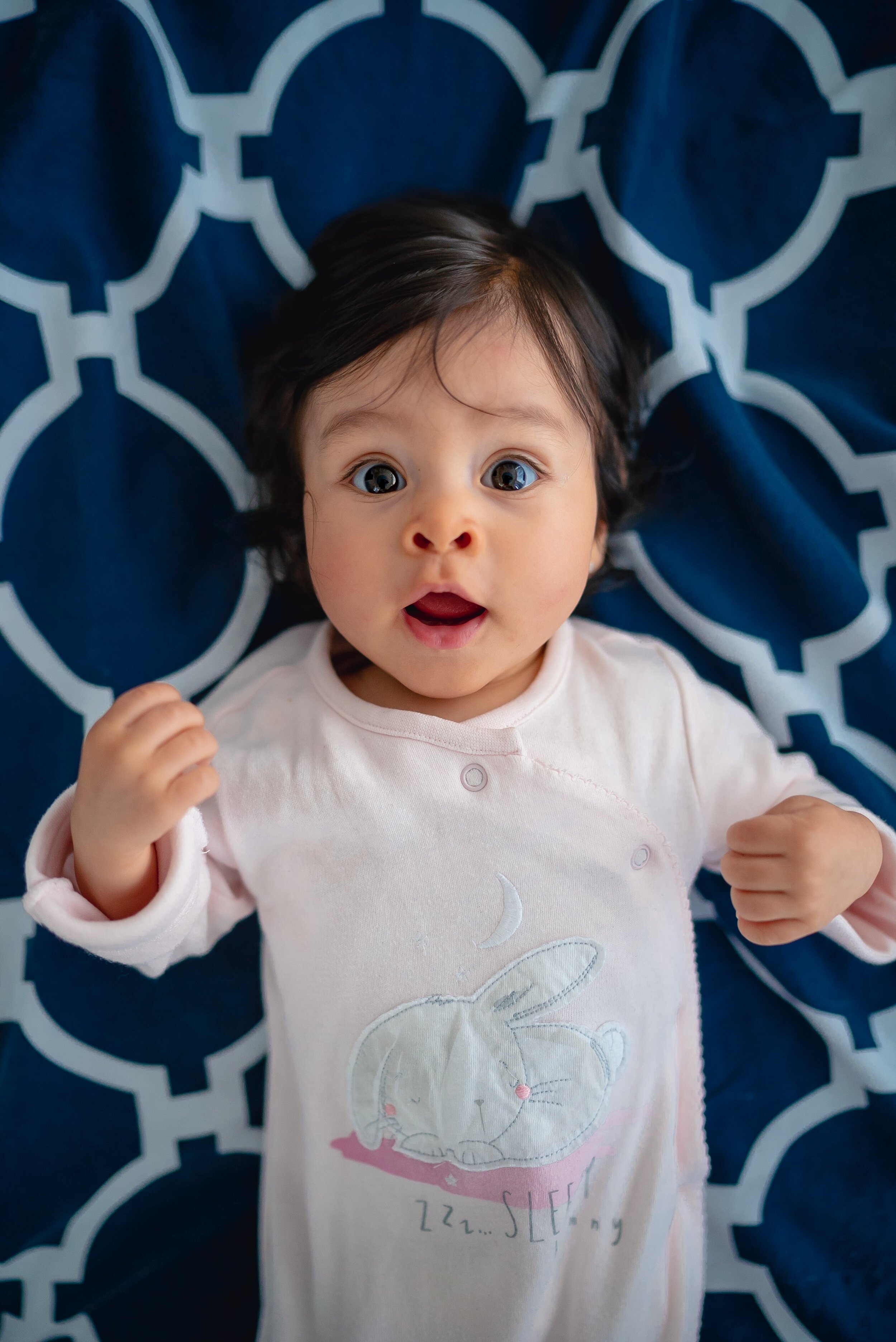  bright-eyed baby laying on back on a blue and white blanket looking at camera 