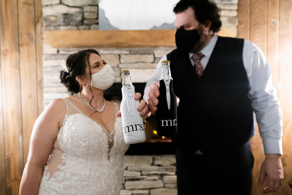 bride and groom wearing facemasks and cheersing corona beers in bottle sleeves