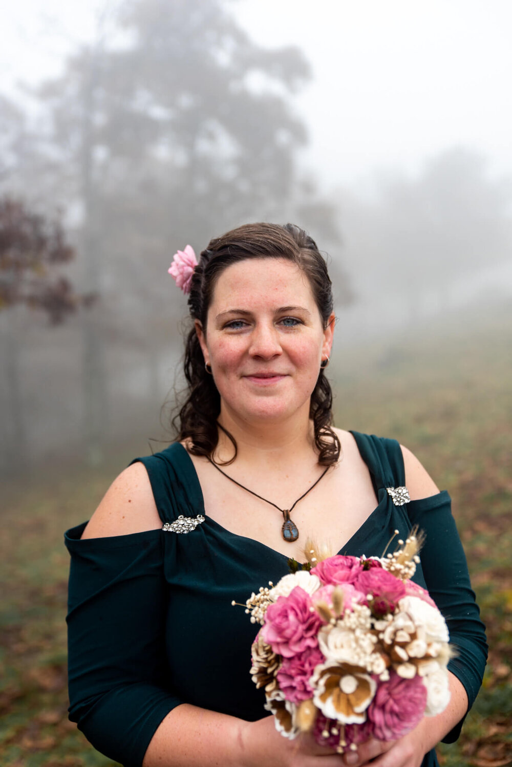 Bride in dark green wedding dress holding a simple pink and white bouquet in a foggy forest