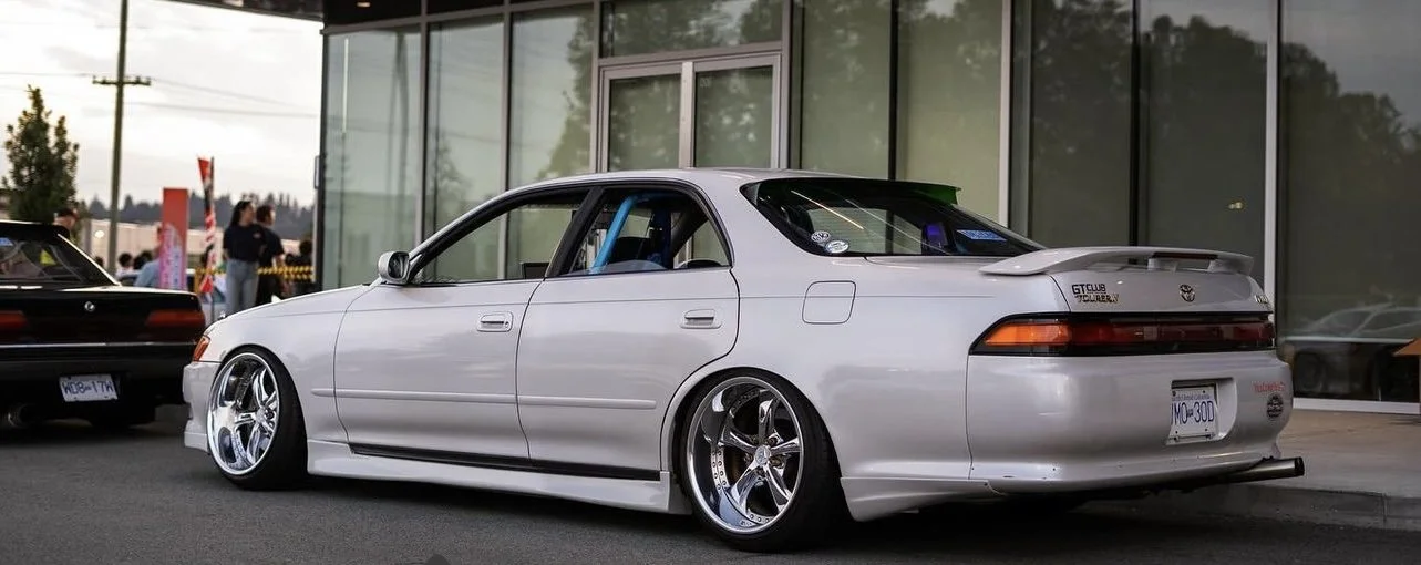 White modified Nissan skyline GTS car with lowered stance, aftermarket wheels, and a custom rear spoiler parked outside a modern glass building. Other vehicles and people are in the background.