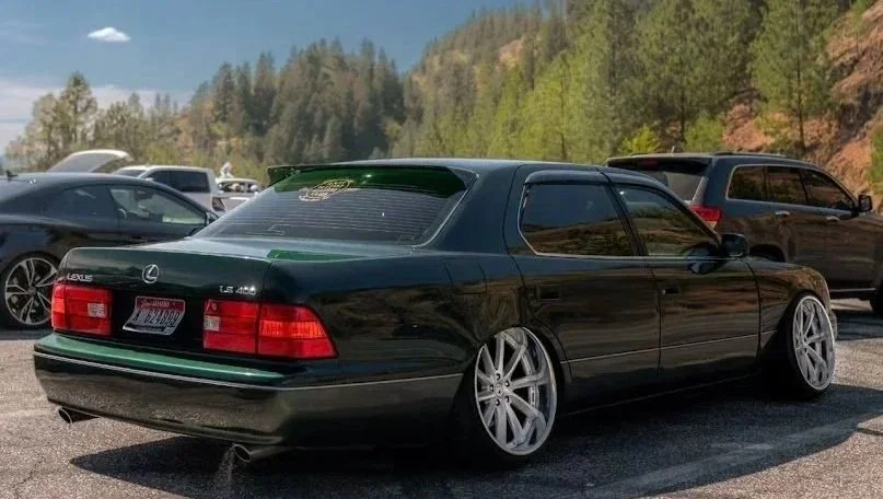 A black Lexus LS 400 with custom large silver wheels parked in a lot, surrounded by other vehicles and trees in the background.