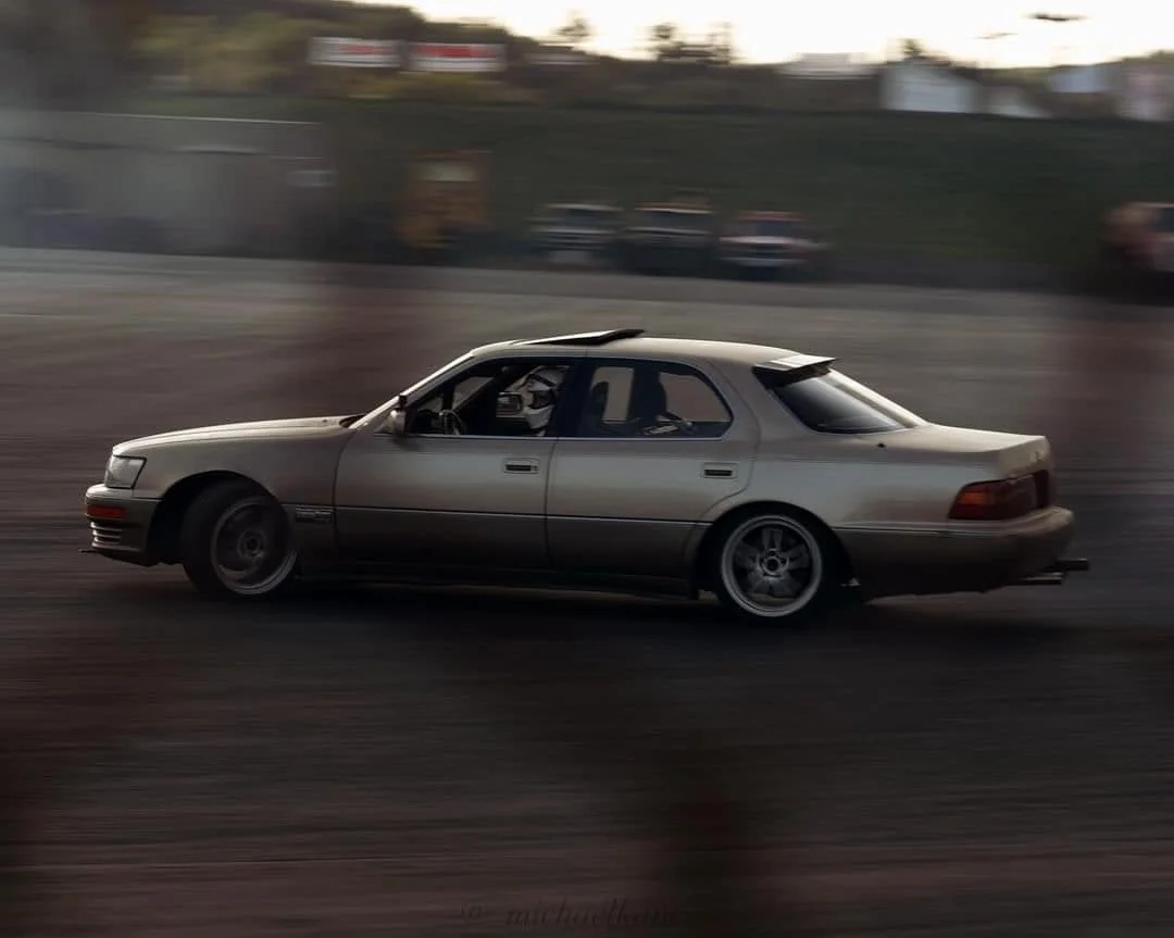 A silver sedan car in motion on a dirt or gravel surface, with a blurred background of other cars and trees, during dusk or dawn.