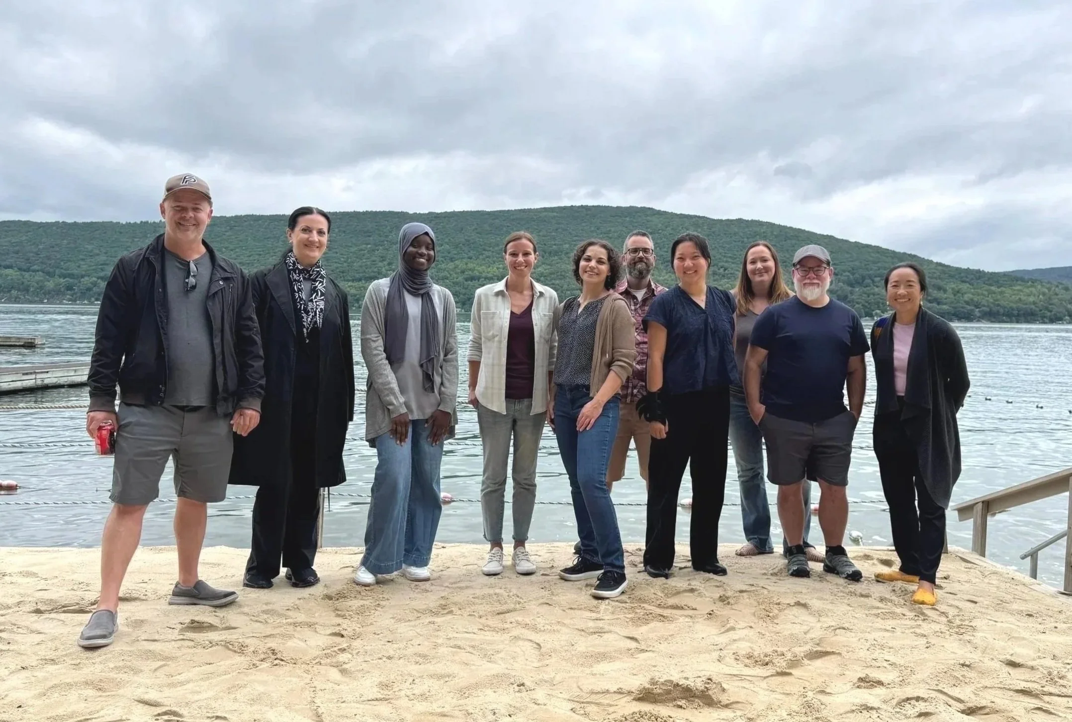 Thumbnail of a group of ten UX team members standing on a sandy beach at Canandaigua lake with hills in the background, overcast sky.