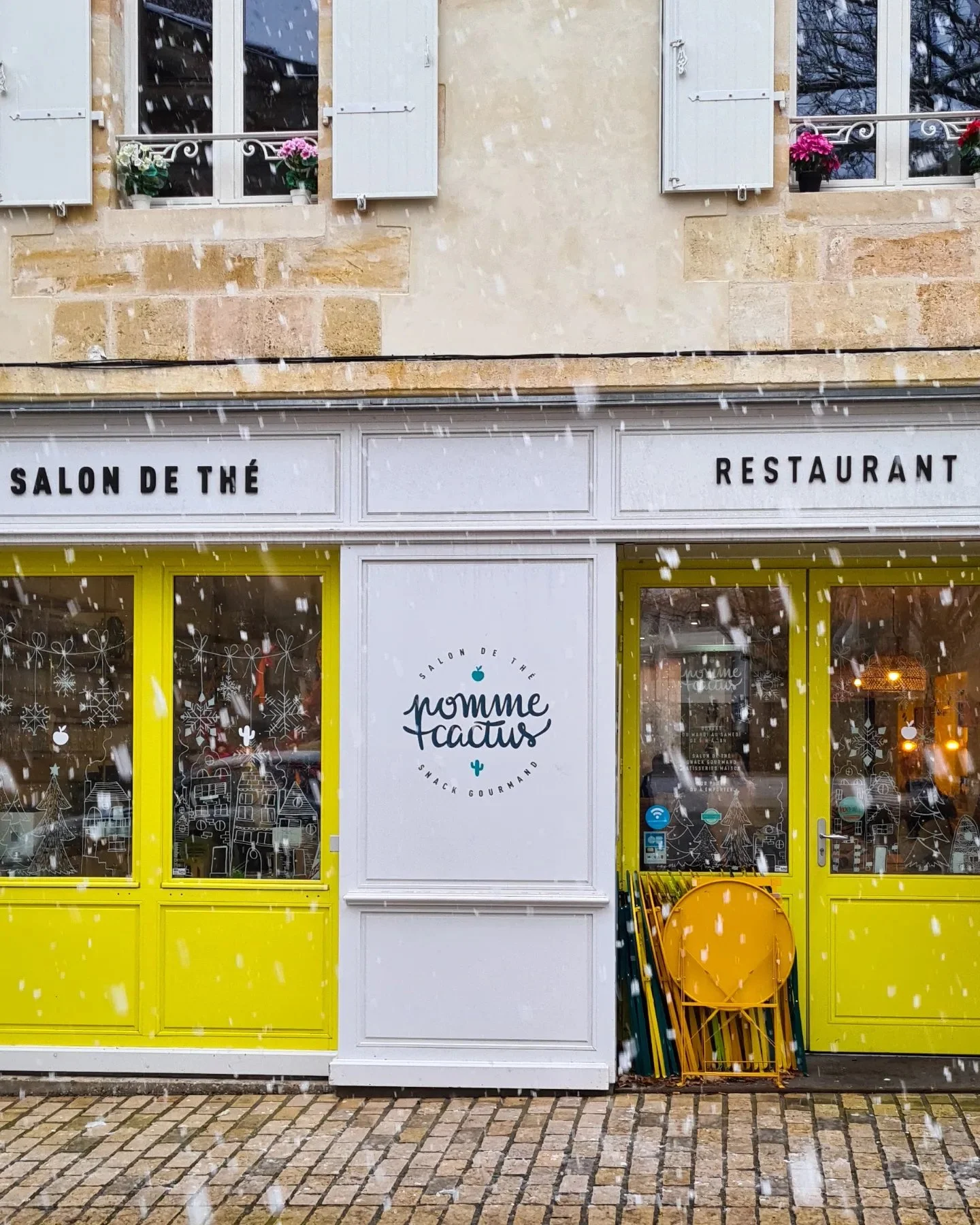 Façade d'un restaurant nommé 'pomme tactus', avec fenêtres décorées de flocons de neige, sur fond de bâtiment en pierre, en hiver avec neige tombante.