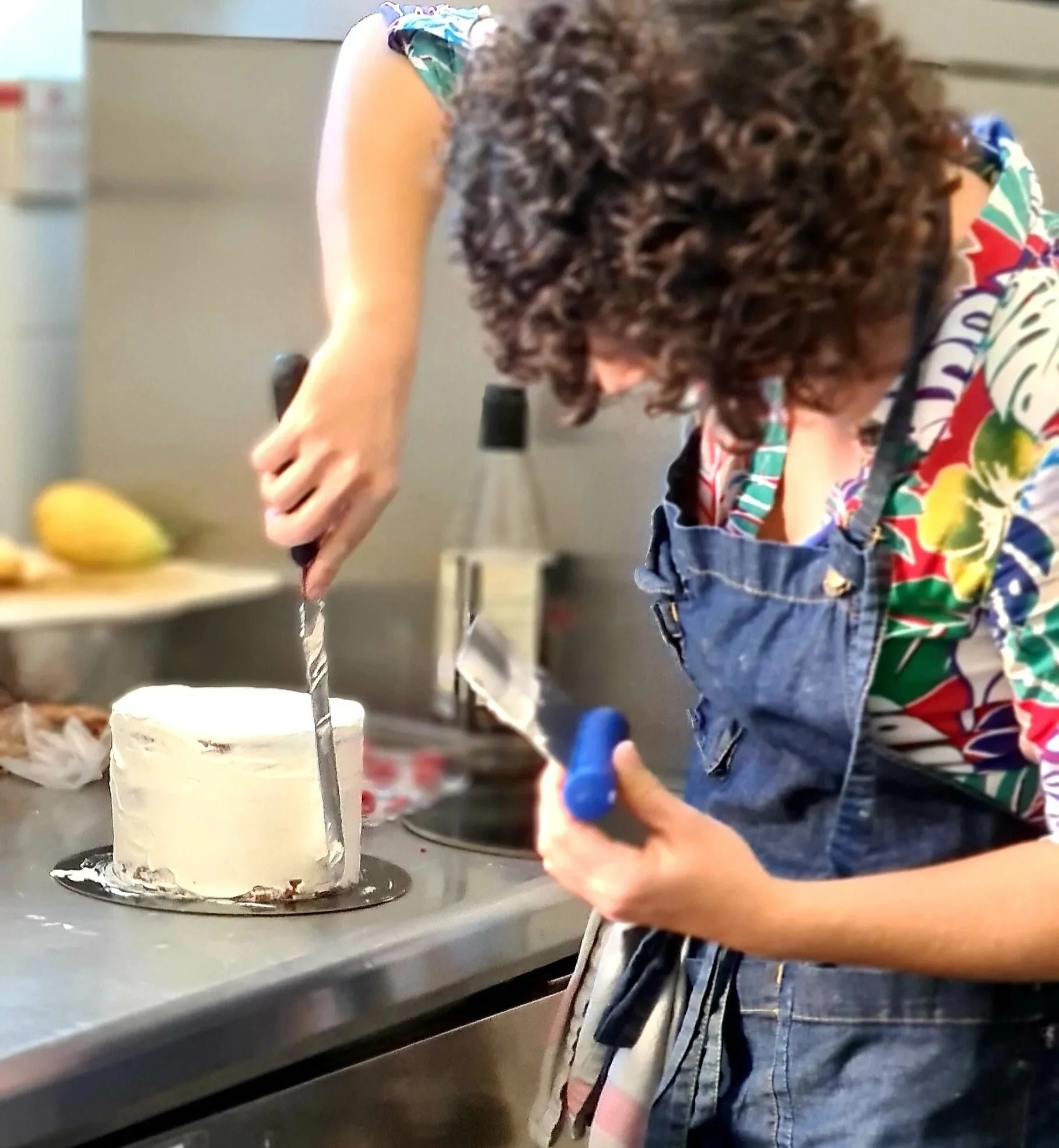 Une femme avec des cheveux bouclés, portant un tablier en jean, coupe un gâteau blanc avec un couteau, dans une cuisine.