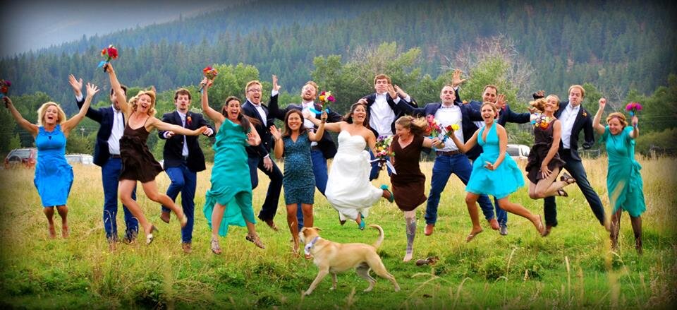 Group of people in formal attire jumping in a grassy field with trees in the background, a bride in the center in a white dress.