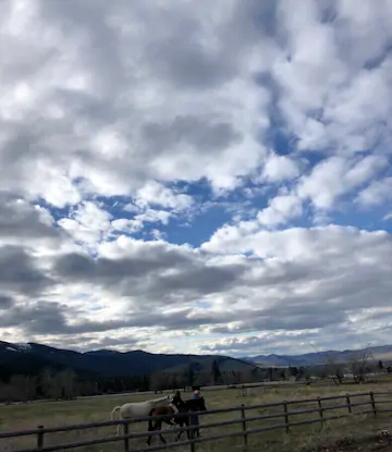 Scenic view of a cloudy sky over a rural landscape with mountains in the background, a wooden fence in the foreground, and people riding horses in a field.