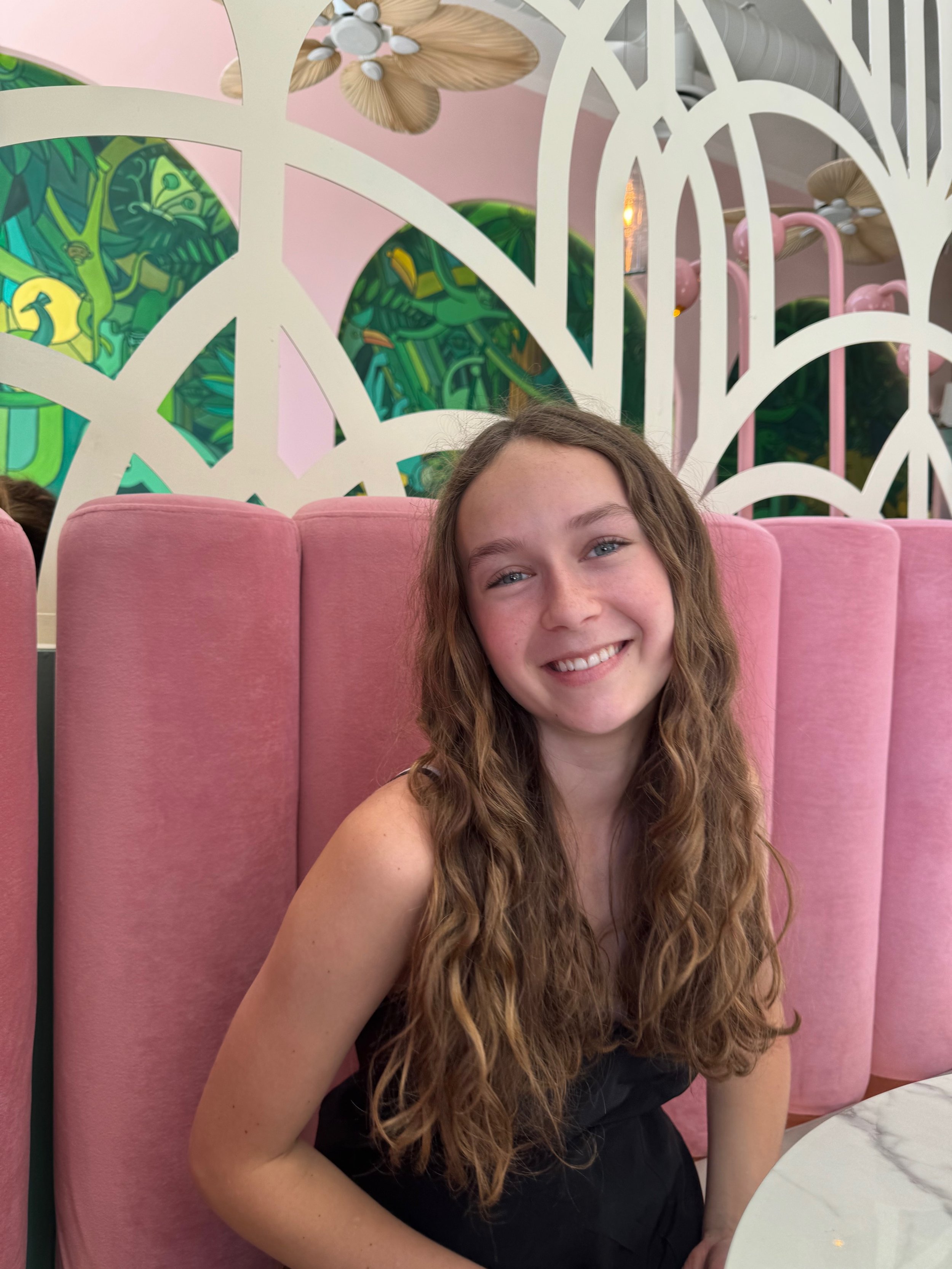A young person with long hair sitting at a table in a restaurant with pink velvet seating and decorative green and white wall design.