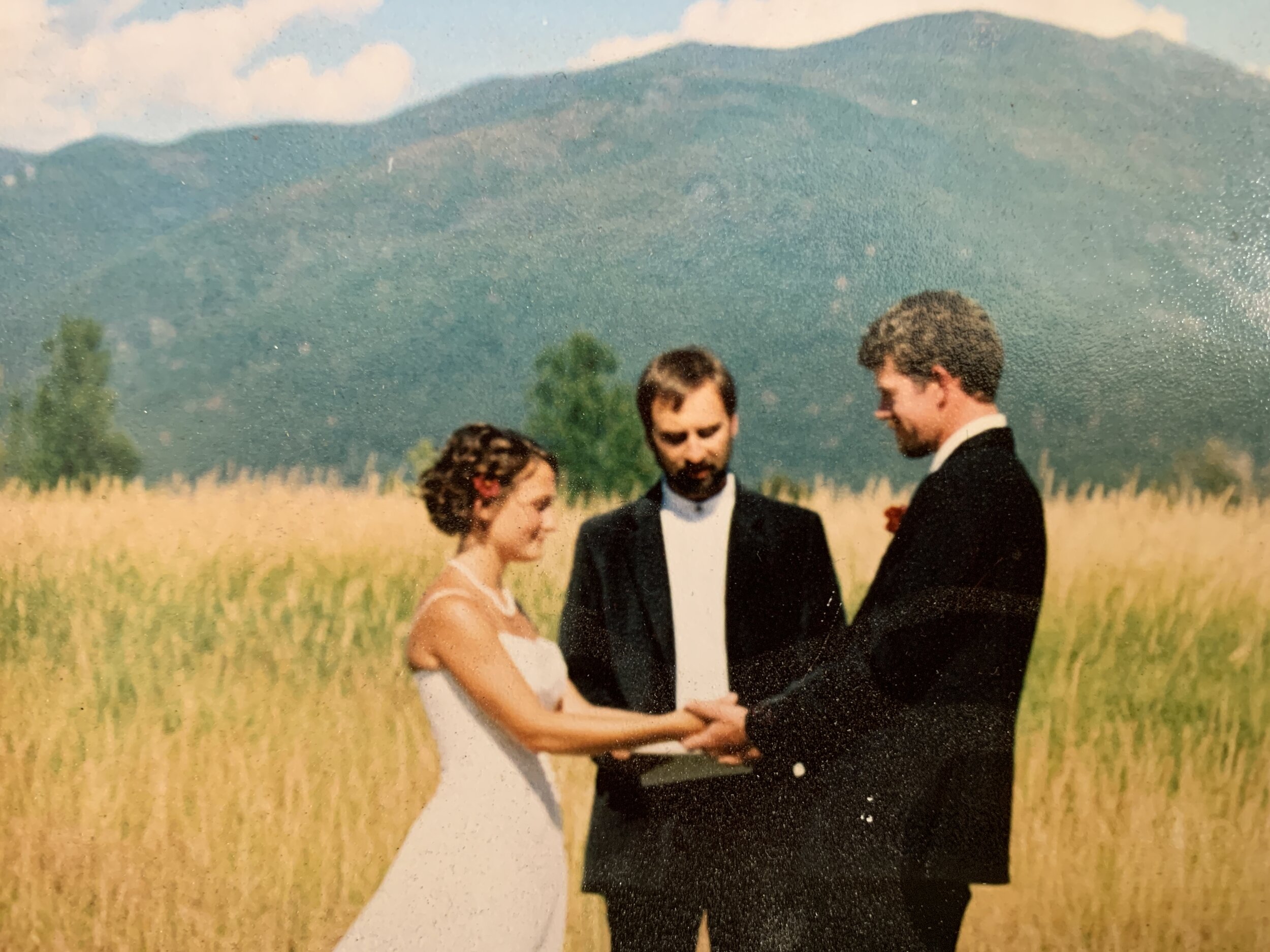Outdoor wedding ceremony with bride and groom holding hands, officiant between them, in a field with mountains in the background.