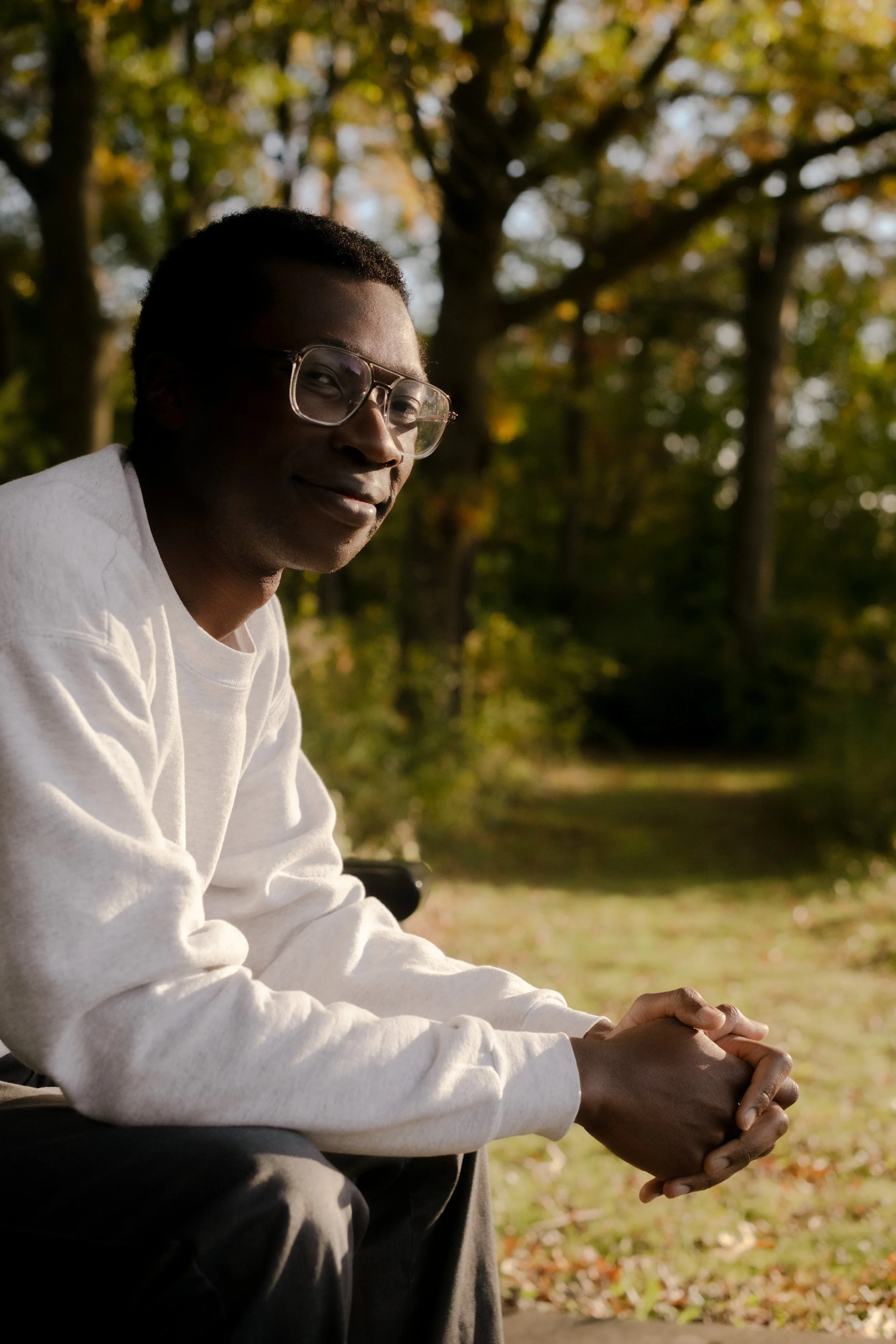 A young man wearing glasses and a white long-sleeve shirt sitting outdoors during autumn, with trees and fallen leaves in the background.