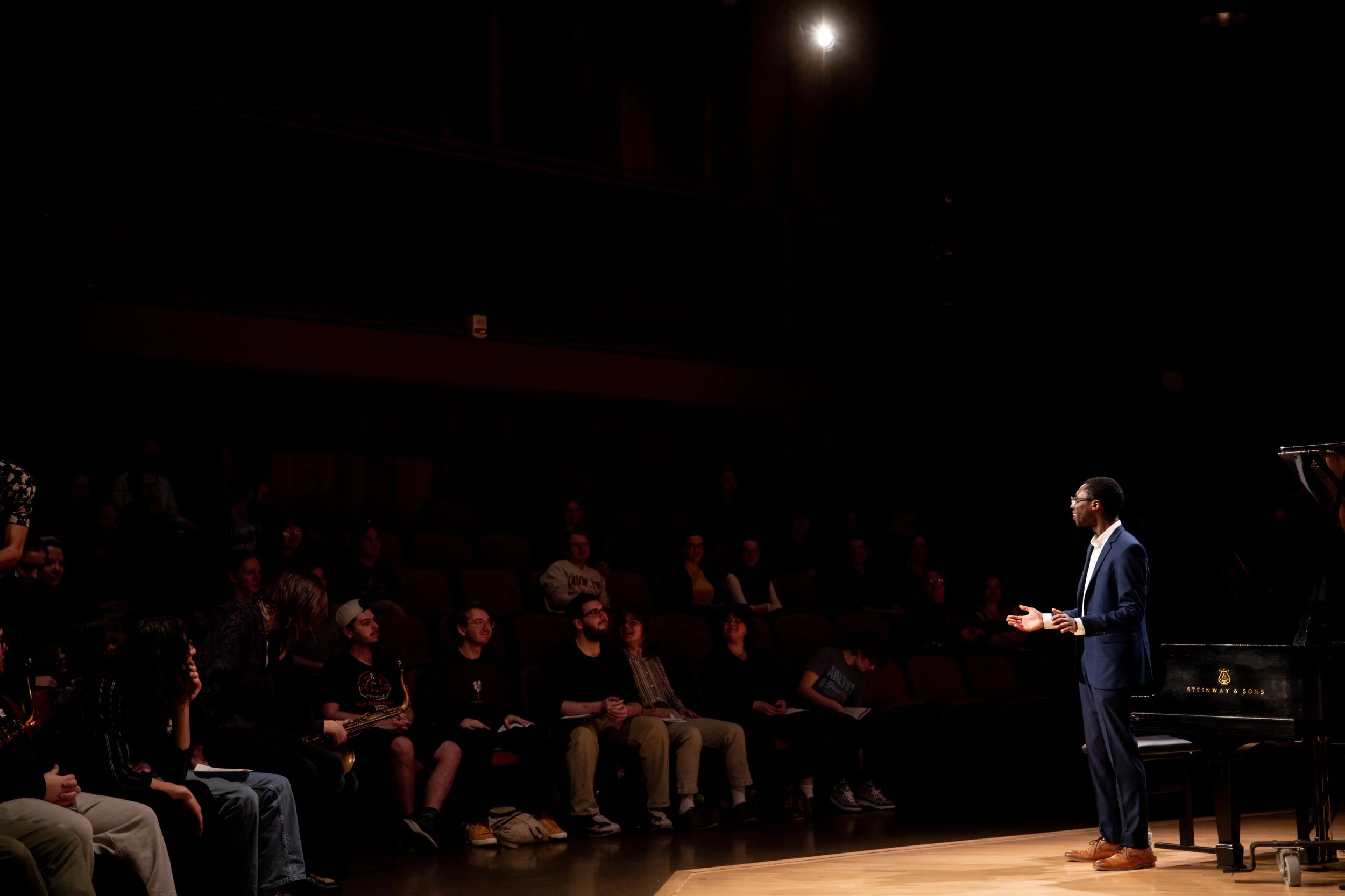 A man in a dark suit and brown shoes is speaking to an audience in a concert hall, with a grand piano on his right. Audience members are seated and focused on him, with some smiling.