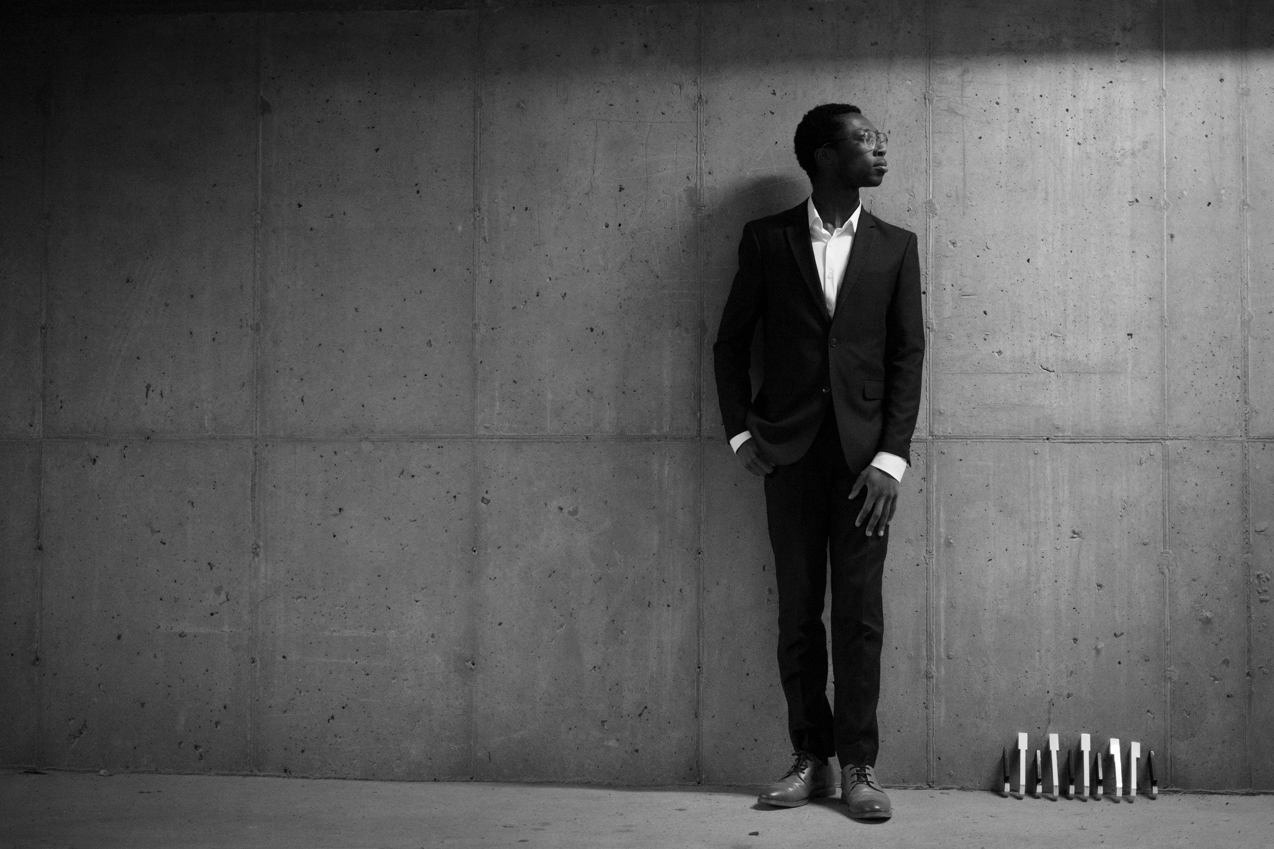 A man in a dark suit and glasses stands against a concrete wall, looking to his right with one hand in his pocket. There are small objects on the floor to his right.