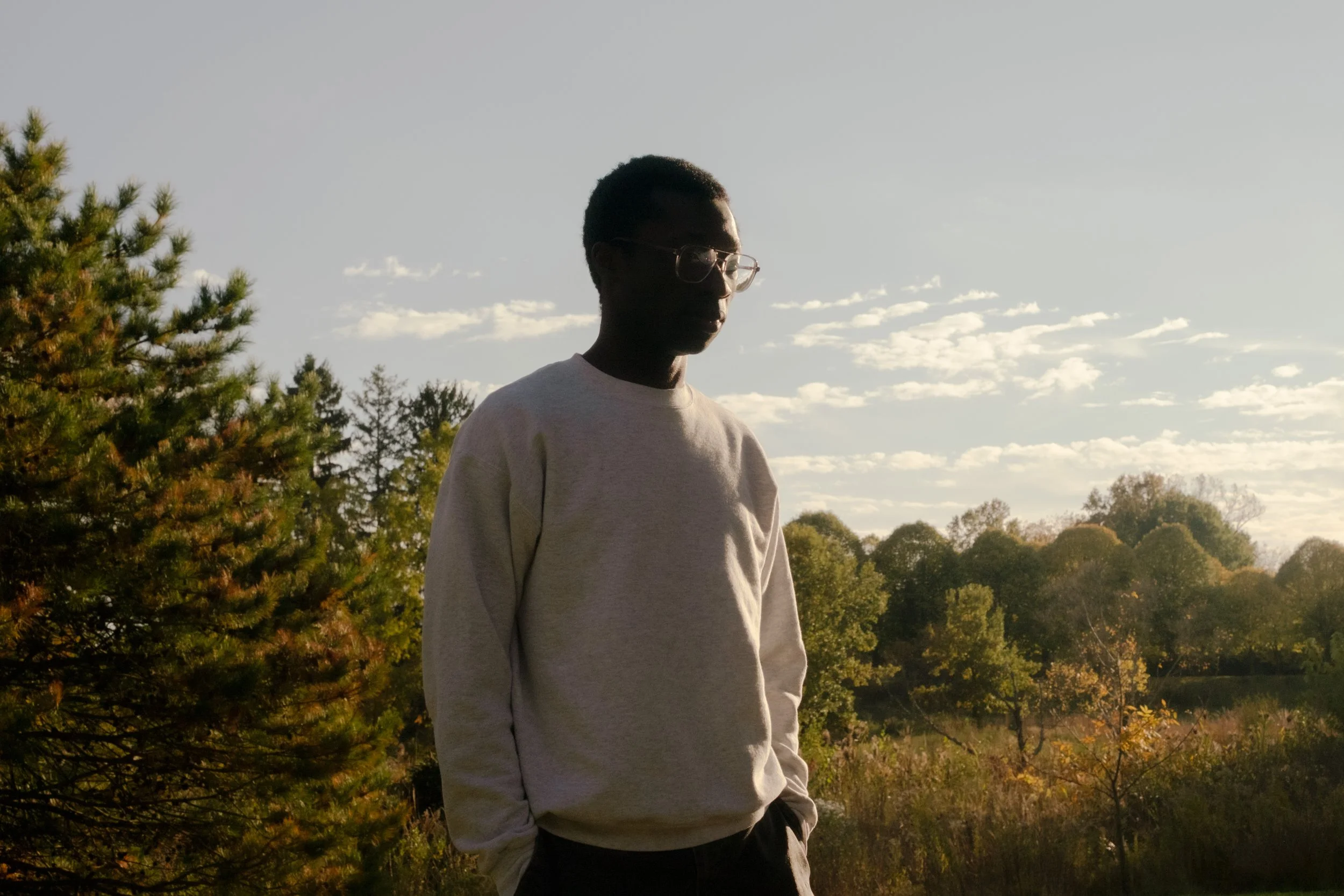 A person with short curly hair and glasses wearing a light-colored sweatshirt, standing outdoors among trees and foliage during sunset.