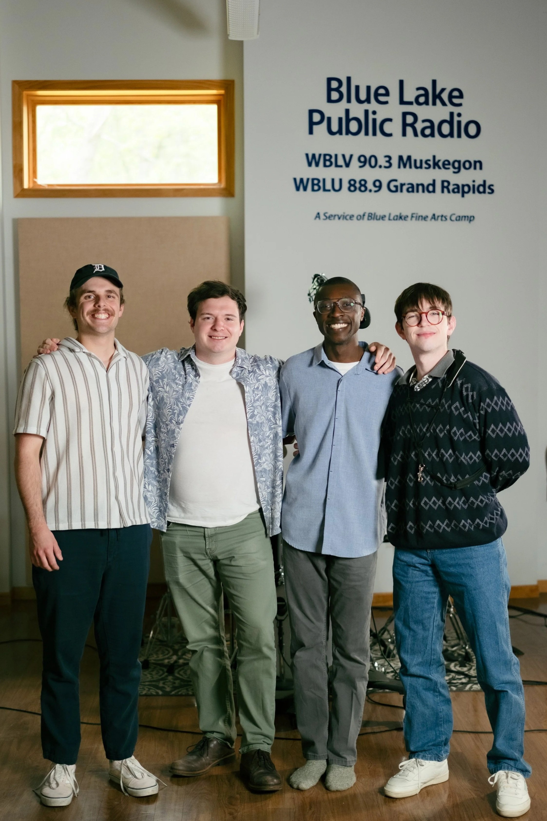 Four young men standing together in a room, smiling at the camera. Signs on the wall indicate it is related to Blue Lake Public Radio, with the stations WBLV 90.3 Muskegon and WBLU 88.9 Grand Rapids. The background includes a window, a beige panel, and a speaker.