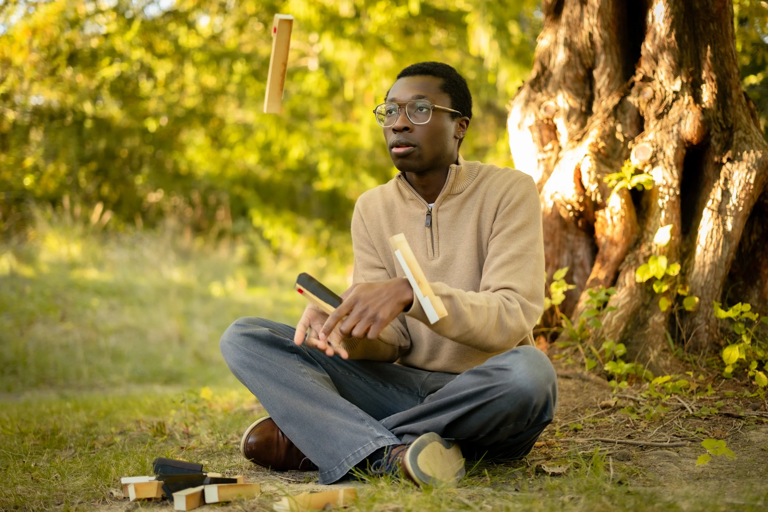 Young man with glasses sitting outdoors under a tree, playing Jenga with wooden blocks, with some blocks on the ground.