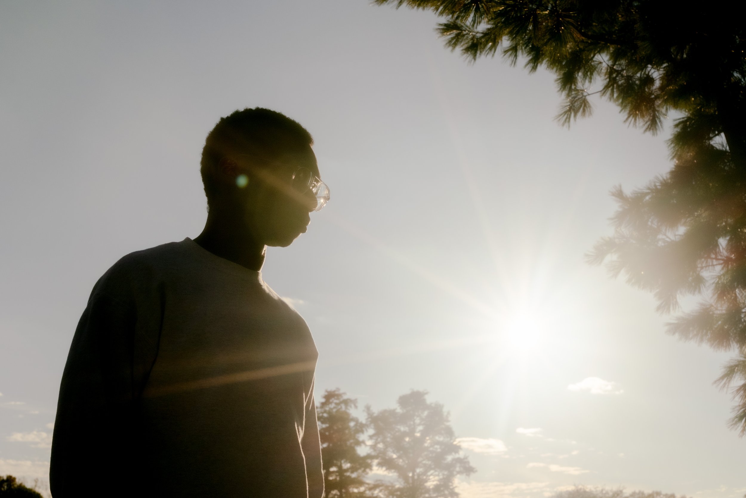 Silhouette of a young man wearing glasses standing outdoors with trees and a bright sky in the background.