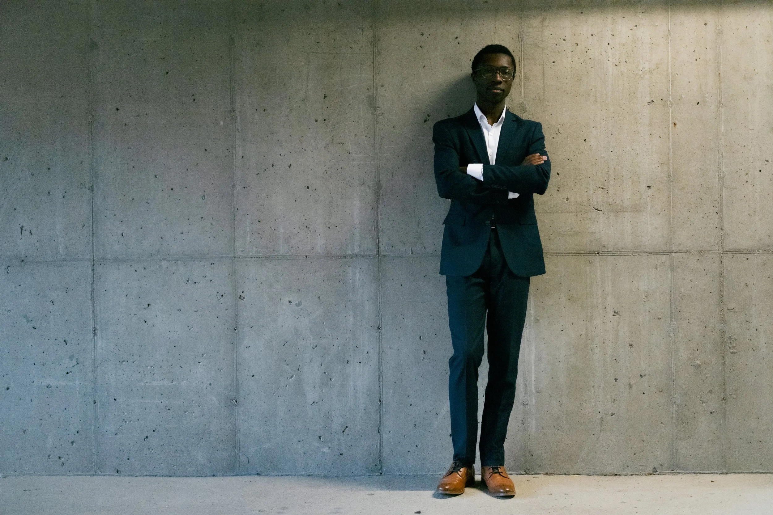 A young man in a suit with glasses, standing with arms crossed against a plain concrete wall.